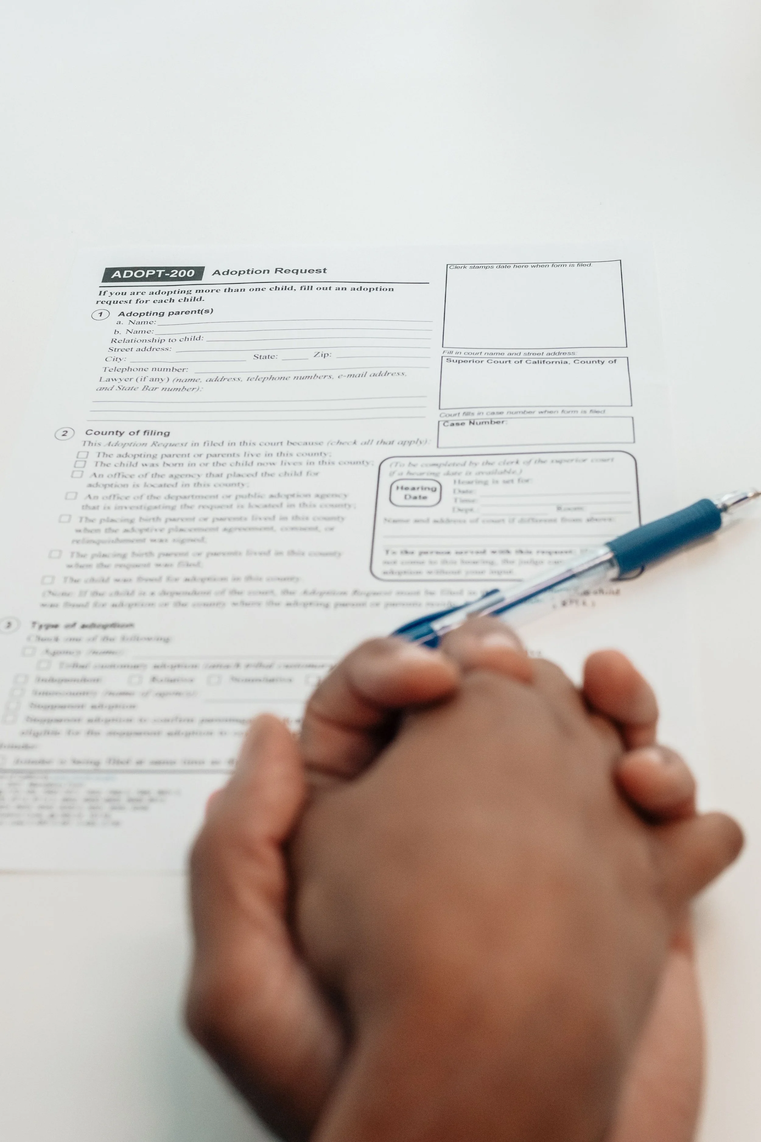 Hands clasped together in front of a printed form labeled 'ADOPT-200 Adoption Request' on a white surface, with a blue pen resting nearby.