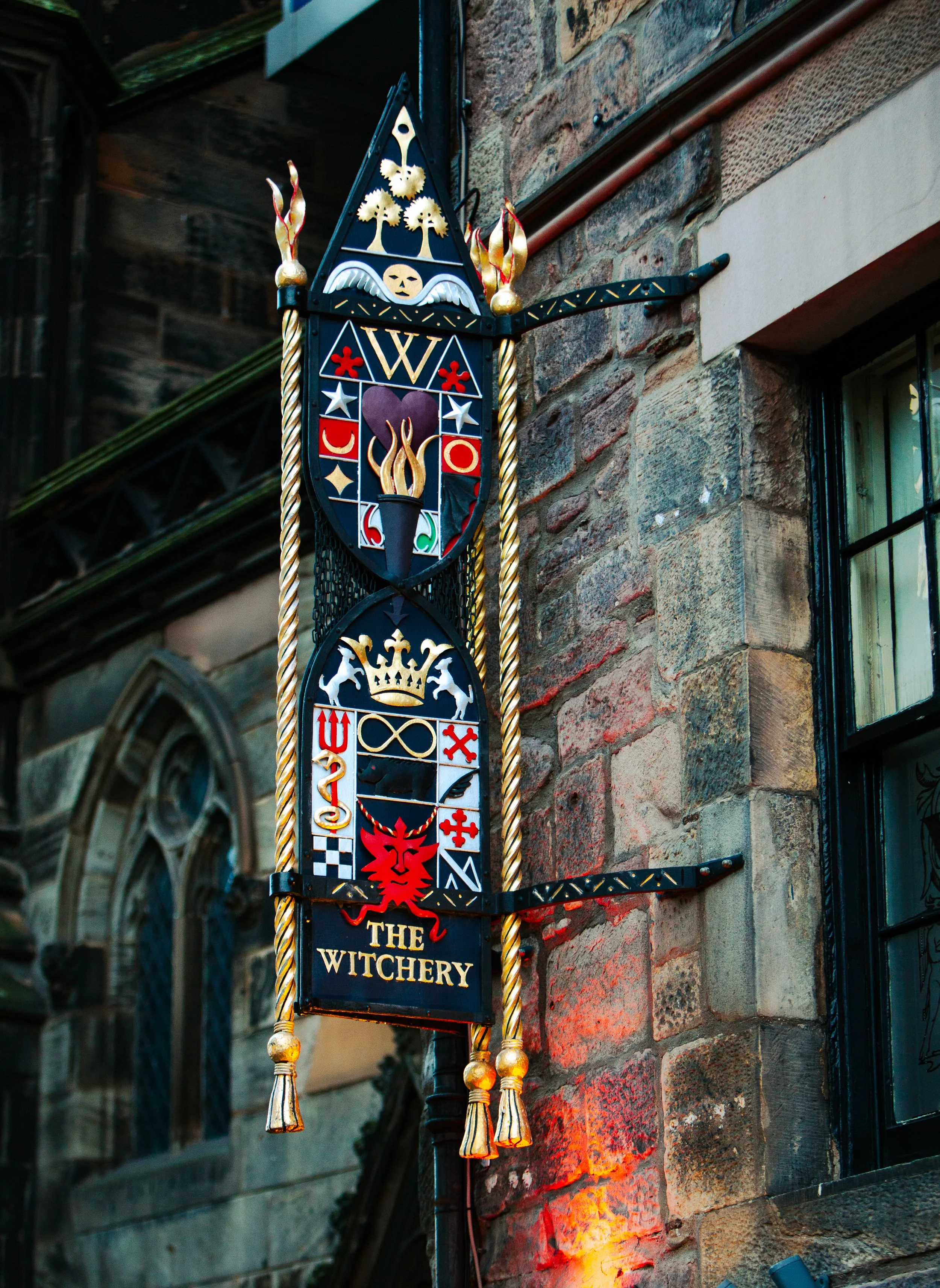 Decorative sign for 'The Witchery' with various symbols, a red lion crest, and a crown, mounted on a brick wall outside a building.
