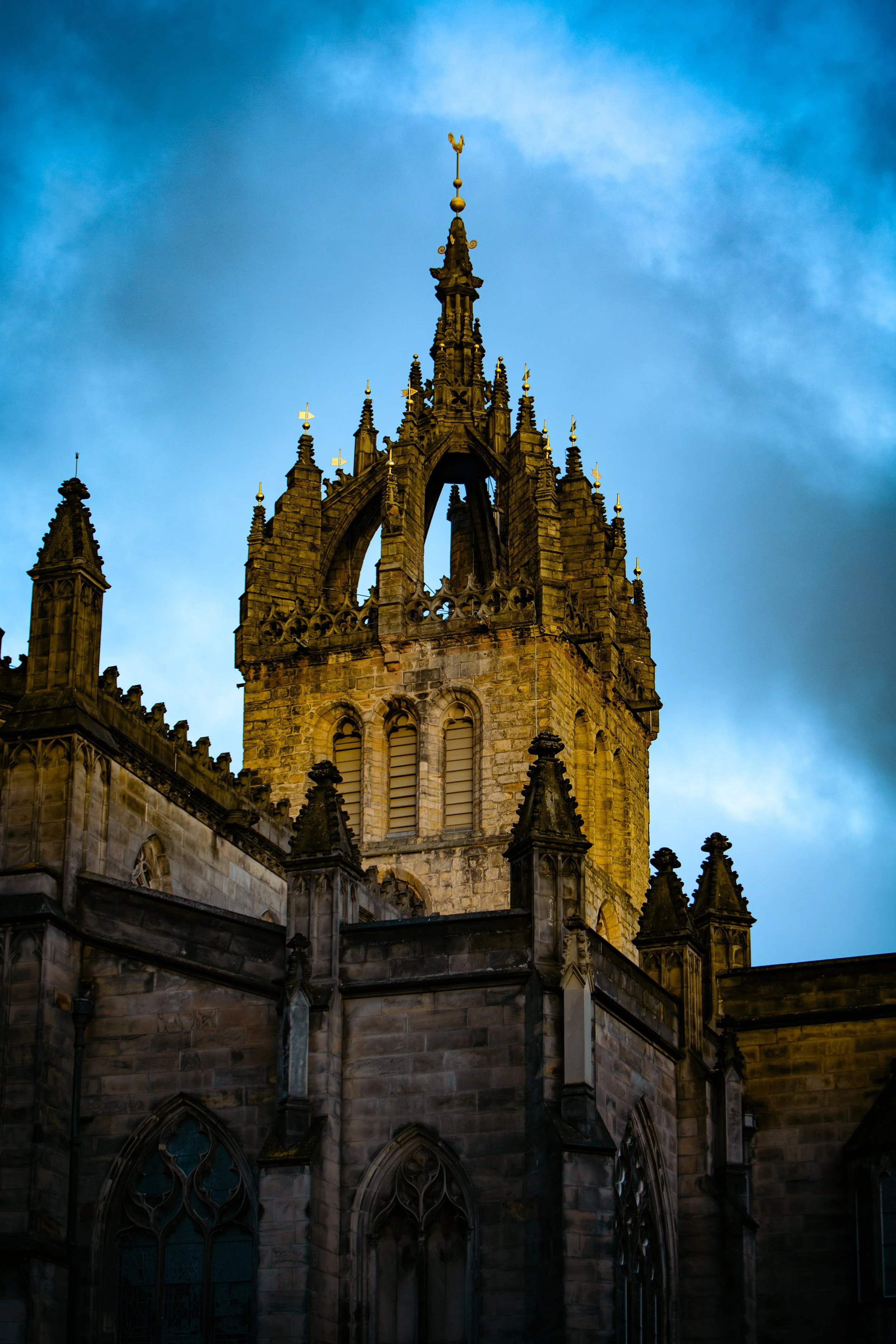 St Giles Catederal Edinburgh with its lantern steeple A Gothic-style church tower made of stone with intricate spires and oculi, set against a blue sky with wispy clouds, illuminated by sunlight.