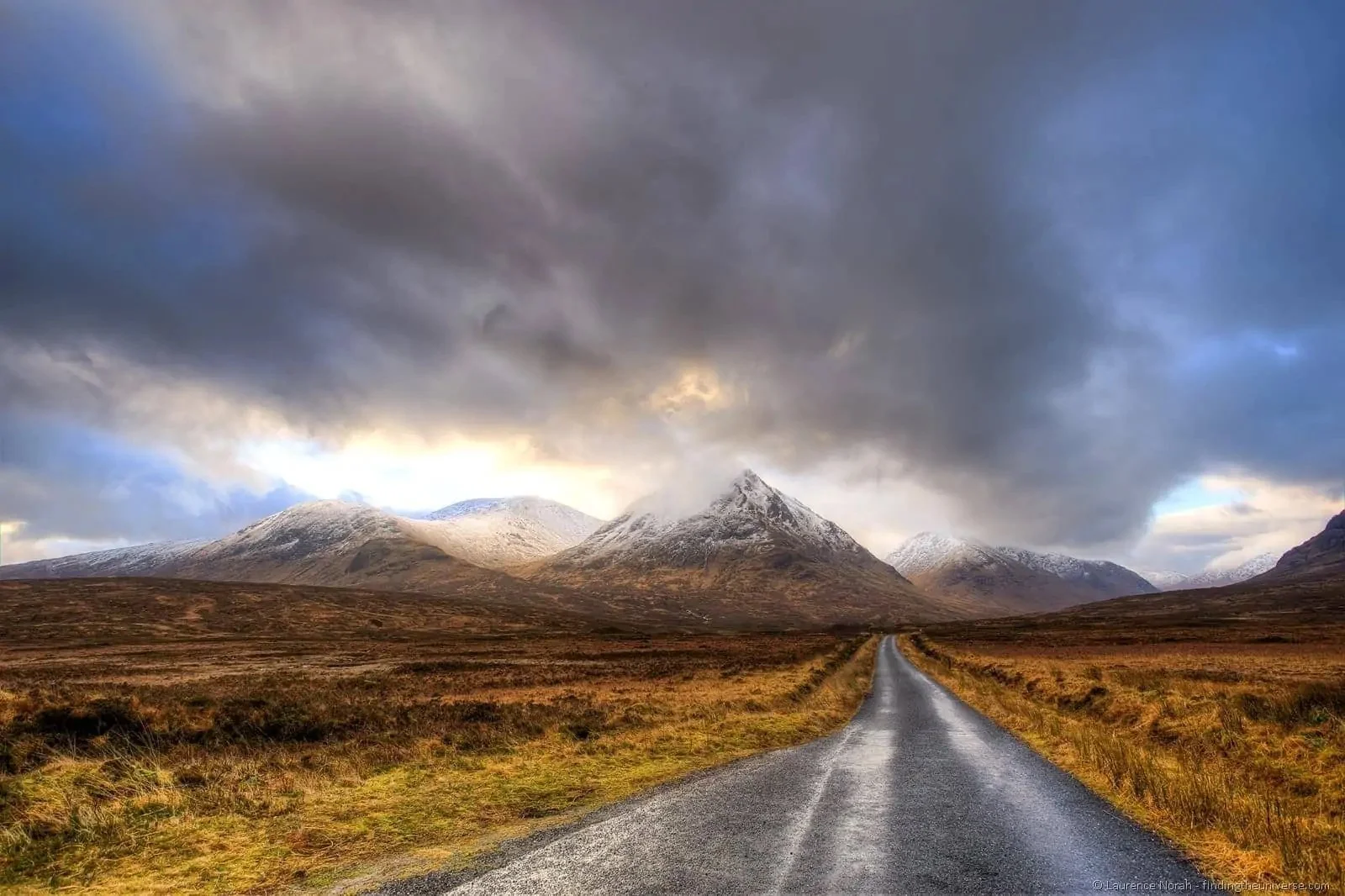 A scenic landscape with a narrow road leading toward snow-capped mountains under dark, cloudy skies.