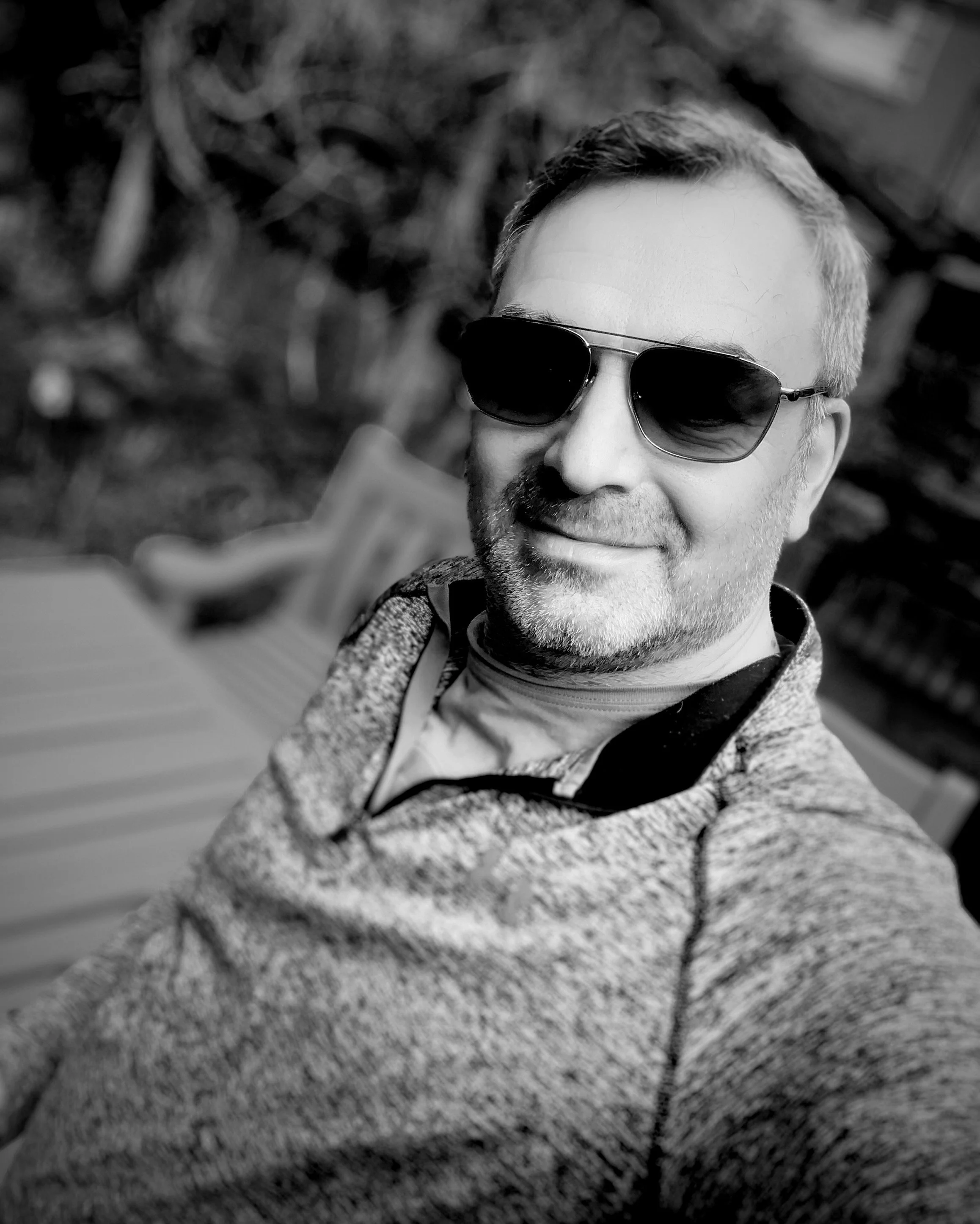 Close-up black and white photo of a man with short hair, beard, wearing dark sunglasses, smiling, with a textured jacket, background blurred with natural elements.