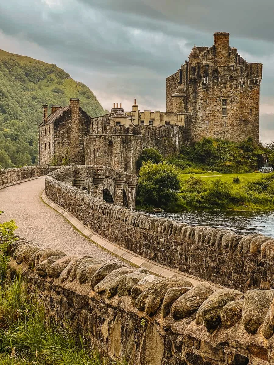 A historic stone castle with a winding bridge leading to it, surrounded by green hills and trees, under a cloudy sky.