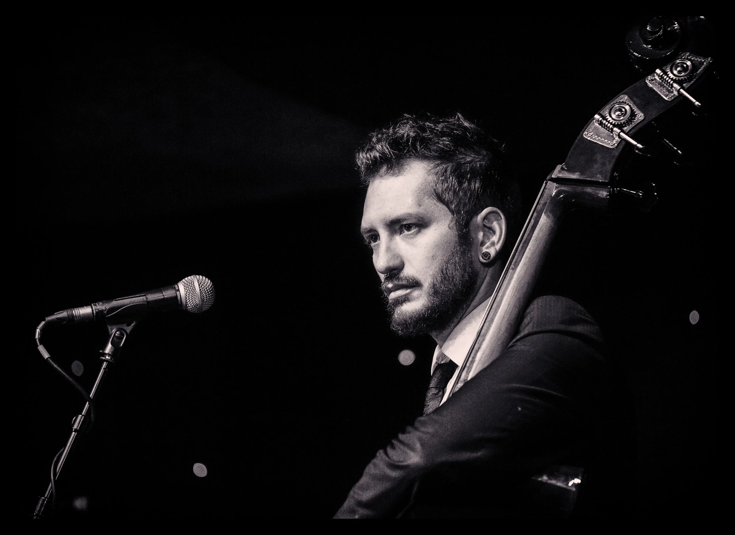 A man with dark hair and a beard, dressed in a suit, playing a guitar on stage with a microphone in front of him, black background.