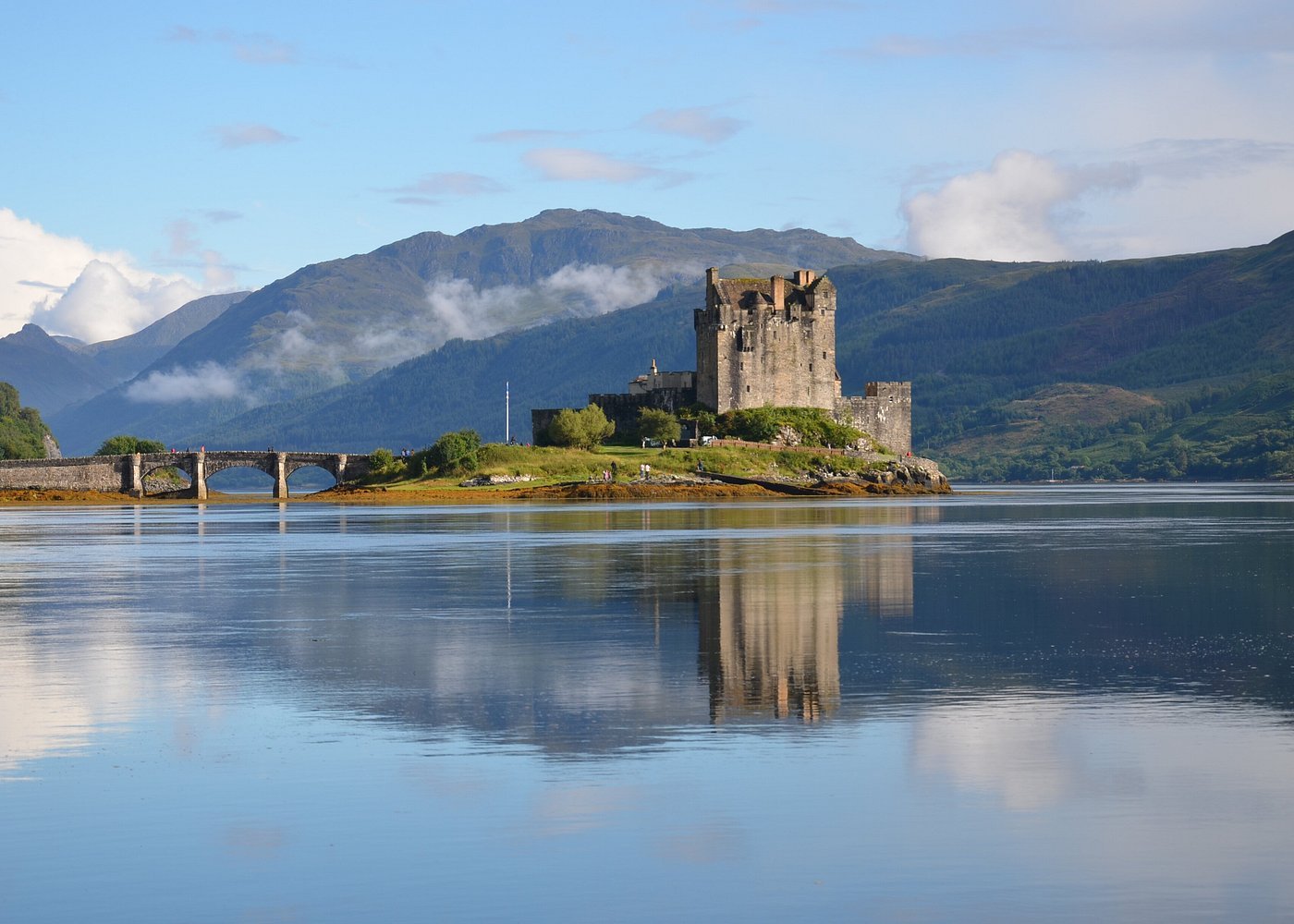 A historic Eilean Donan castle situated on a small island, connected by a stone bridge, with mountains and a partly cloudy sky in the background, reflected in a calm body of water.