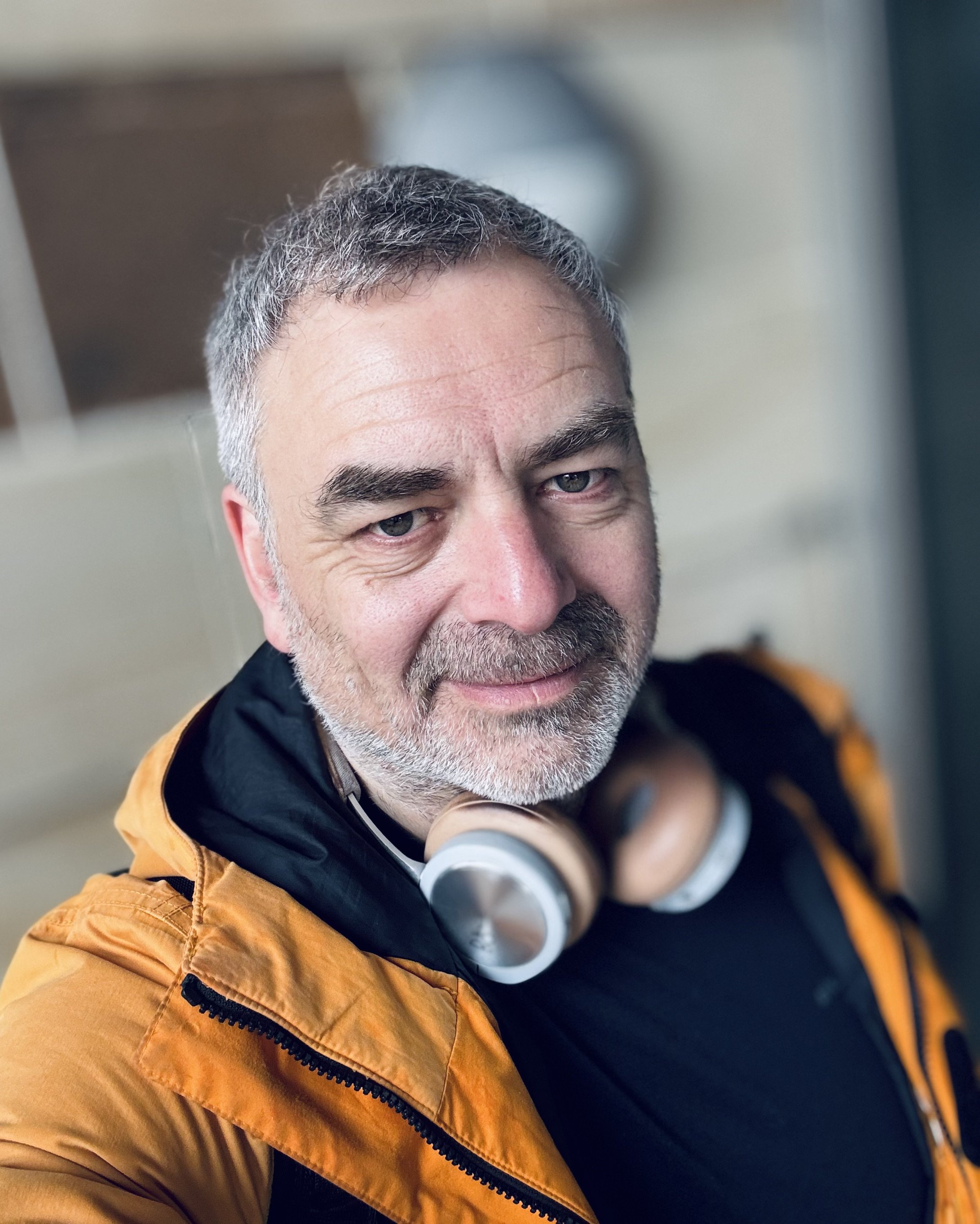 Close-up of a man with gray hair and beard, wearing a black and orange jacket with a zipper, and a gray pair of headphones around his neck.
