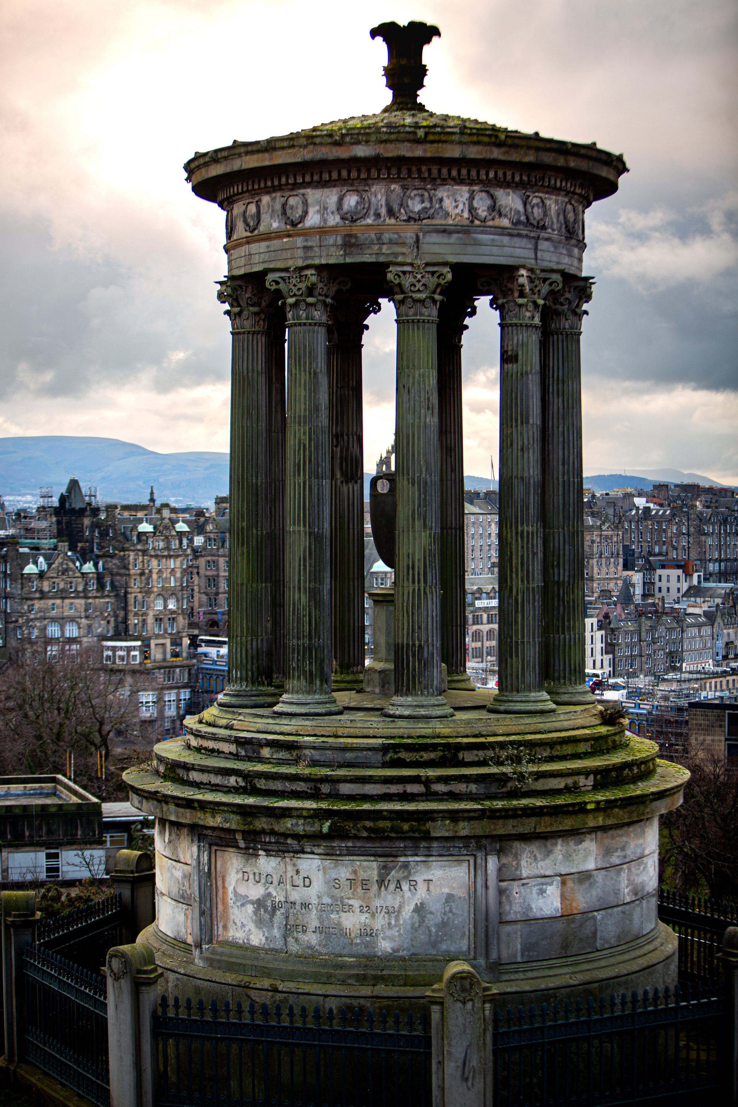 A stone monument with several tall, fluted columns supporting a circular entablature, surrounded by a black wrought iron fence, with buildings and mountains in the distance under cloudy skies. Calton Hill Edinburgh