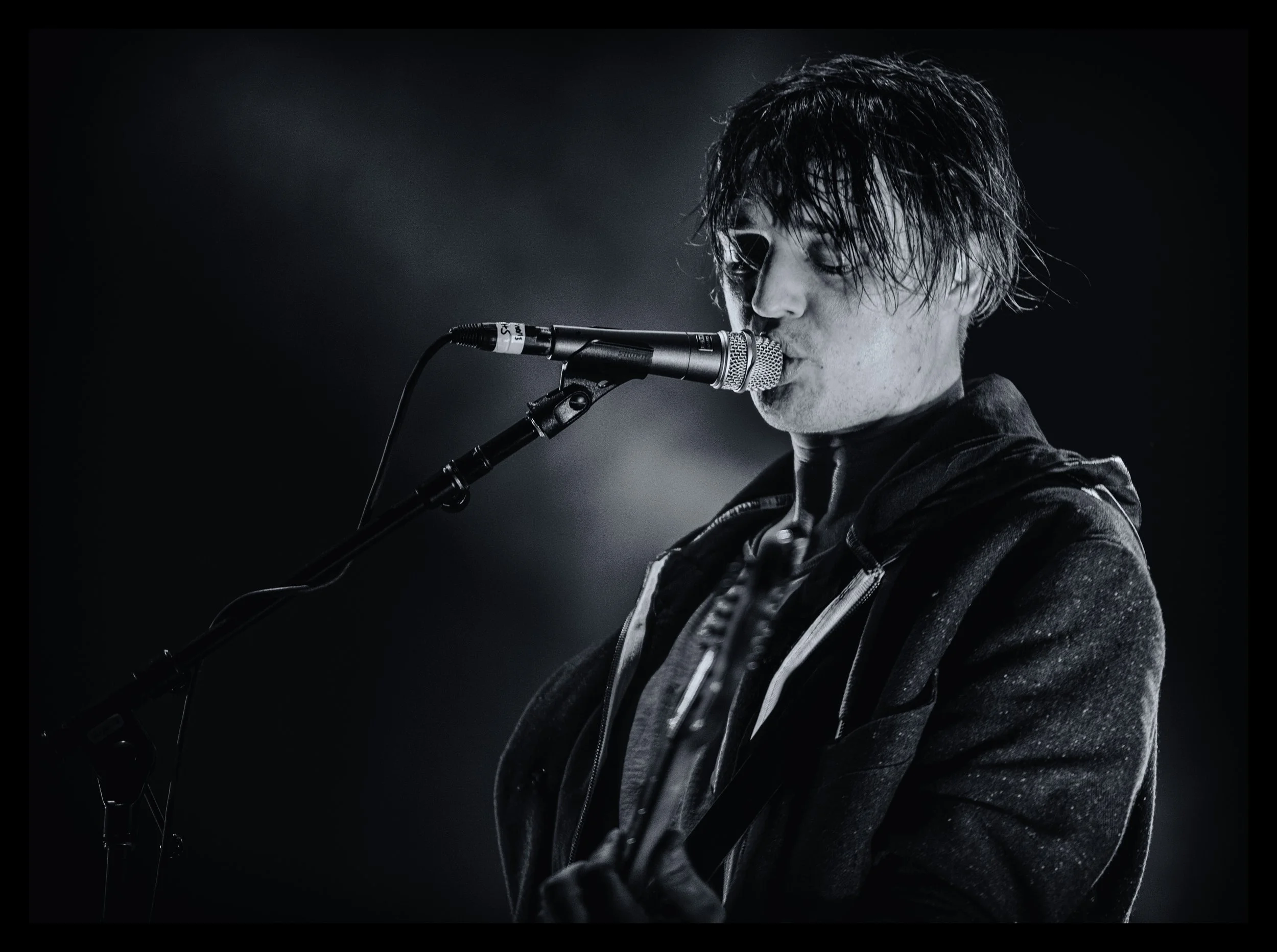 Black and white photo of a male musician singing into a microphone, with wet hair and wearing a dark jacket.