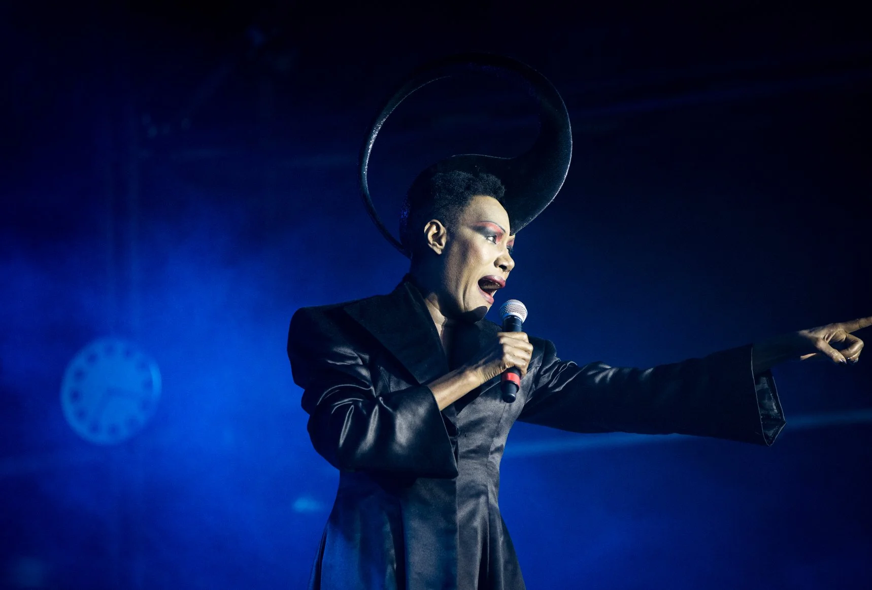 Performer wearing bold makeup and a large headpiece, holding a microphone and pointing, on stage with blue lighting.