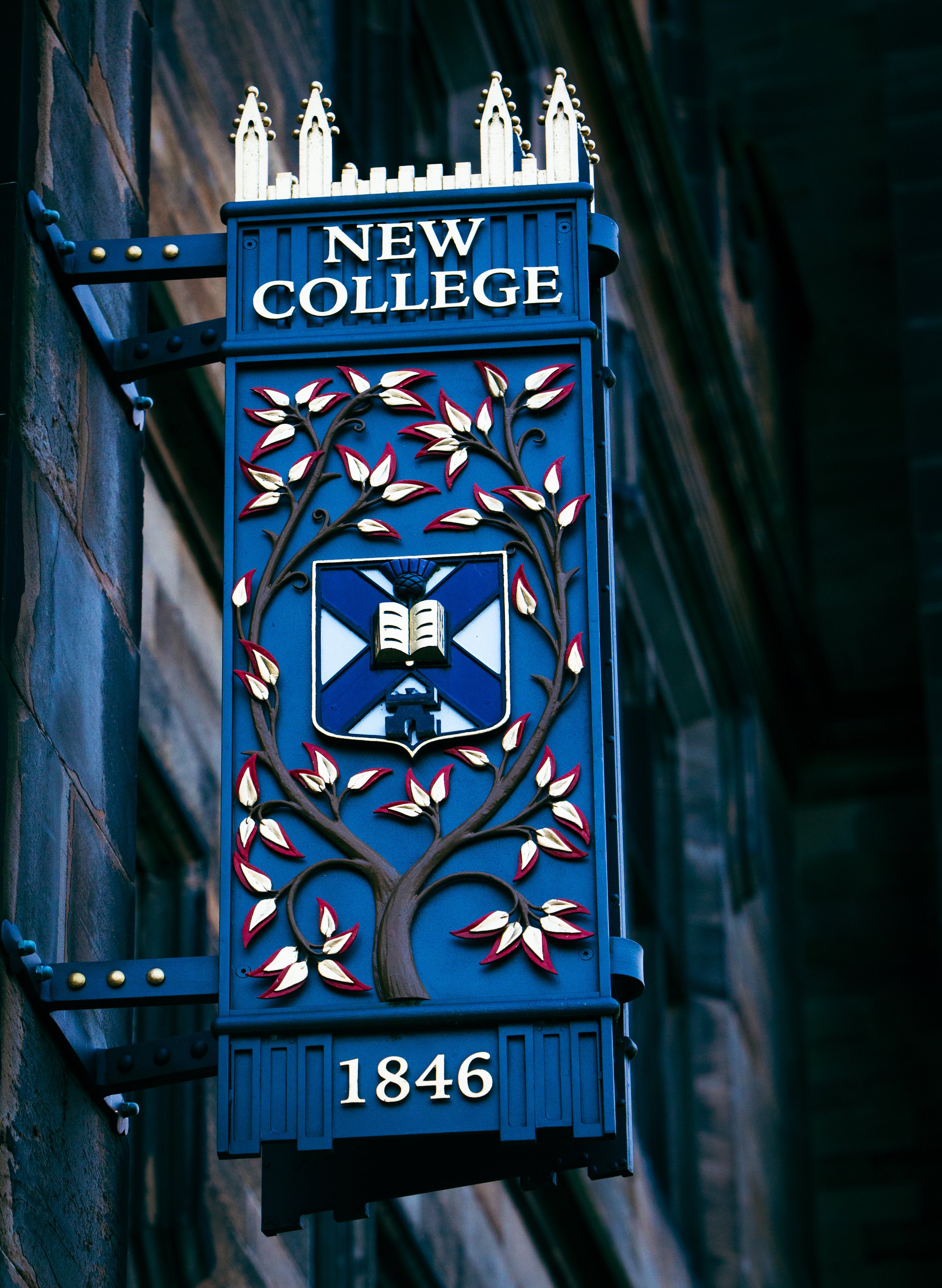 A decorative sign for New College mounted on a brick building wall, featuring a tree with leaves and an open book emblem, and the year 1846 at the bottom.