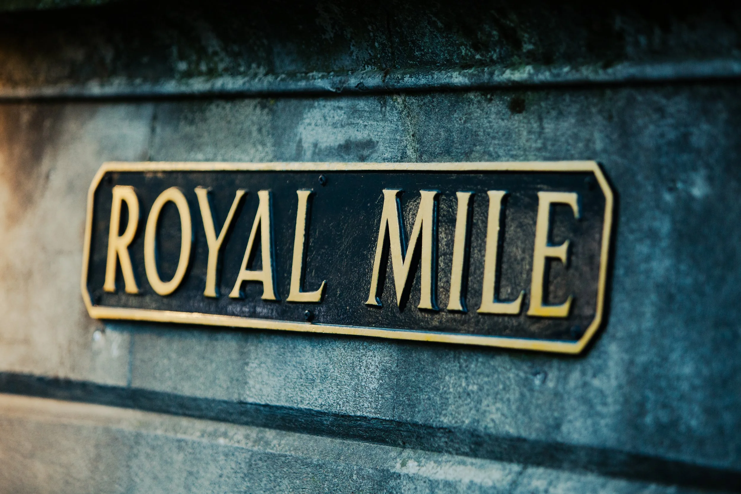Close-up of a brass-colored street sign reading 'Royal Mile' attached to a textured gray stone wall.