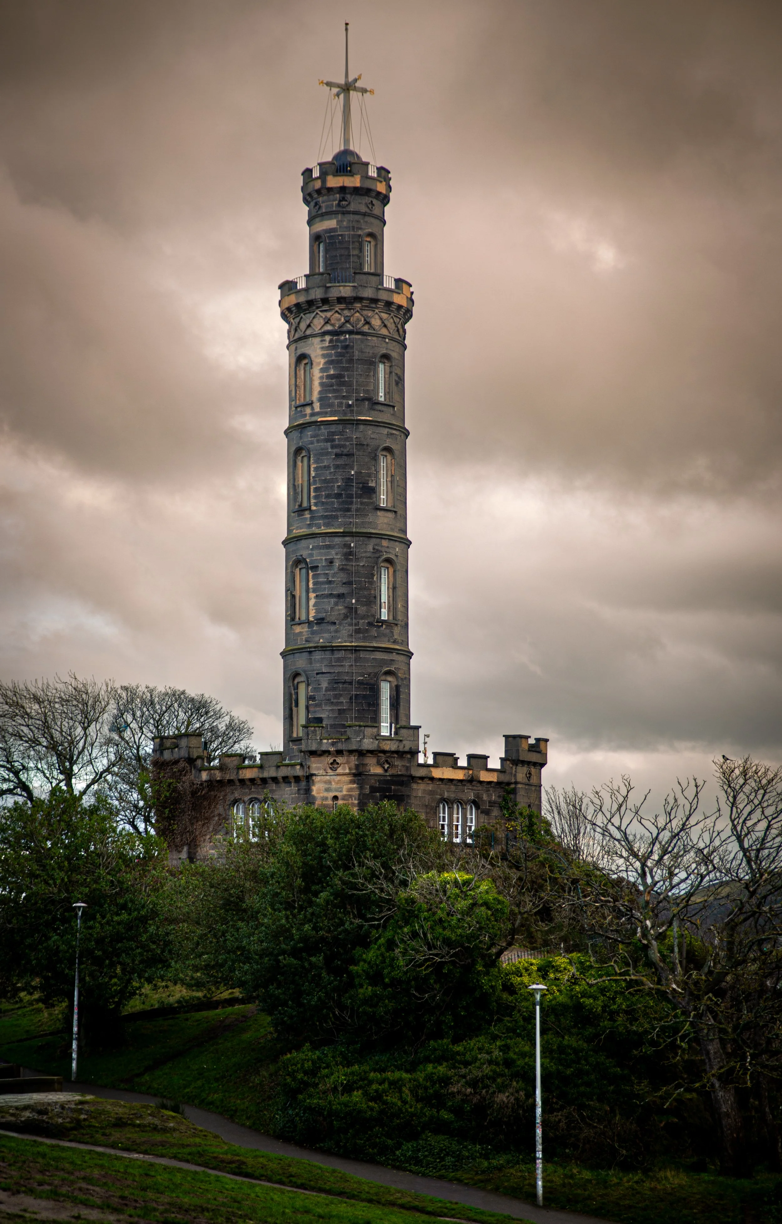 A tall, dark stone tower with a circular shape and narrow arched windows, surrounded by trees and a park with winding paths and street lamps, under a cloudy sky.