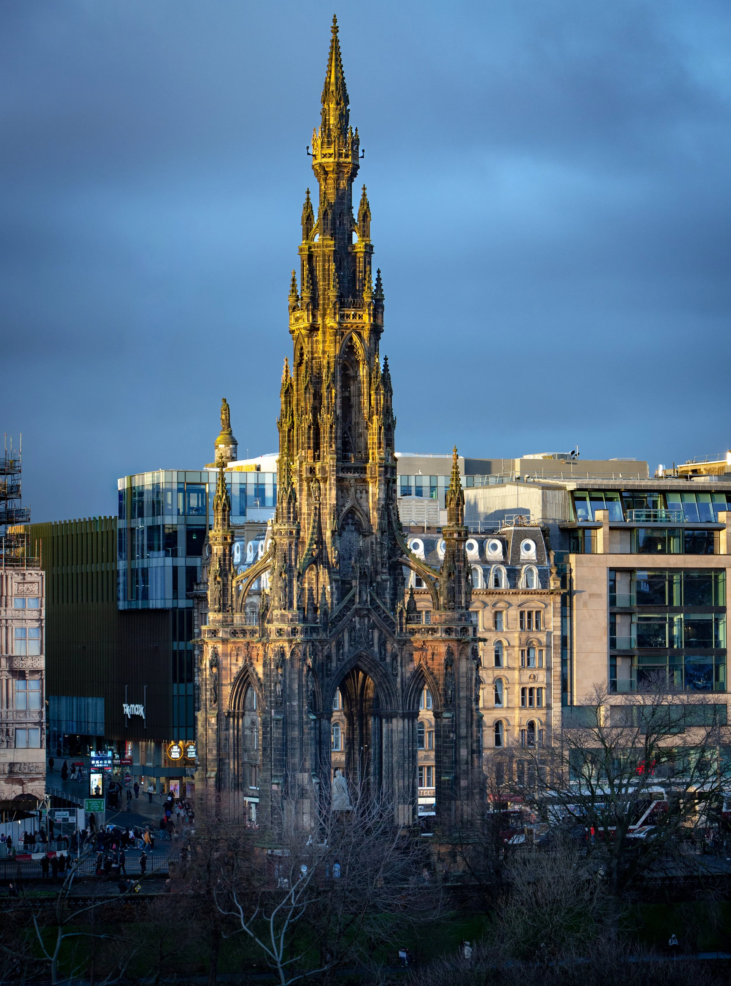 A tall, ornate Gothic-style church tower illuminated with golden light, contrasting against a dark, cloudy sky in an urban setting. Modern buildings and city streets with people are visible at the base.