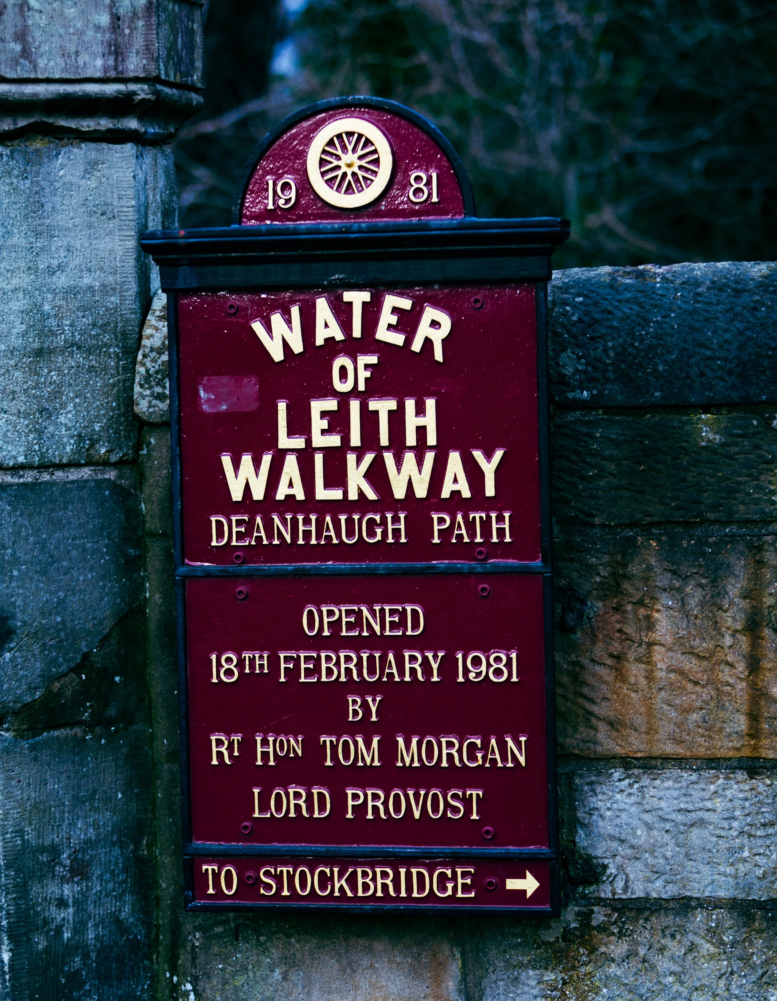 A red and black informational sign with golden text. The top part shows a decorative wheel symbol with the year 1981 on either side. The main text reads 'Water of Leith Walkway' and 'Deanhaugh Path'. Additional text mentions the sign being opened on 18th February 1981 by Rt Hon Tom Morgan, Lord Provost, and indicates the pathway leads to Stockbridge with an arrow.