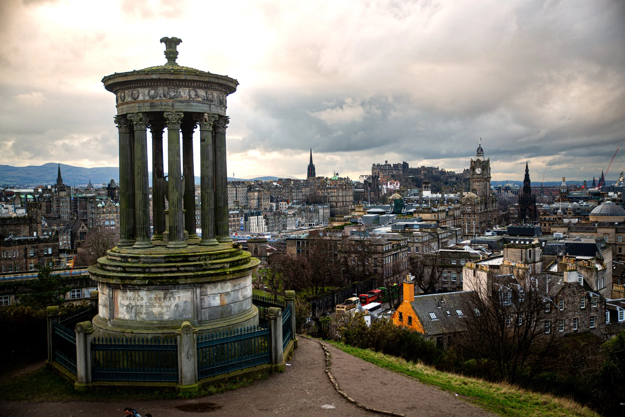View of Edinburgh, Scotland, skyline with historical buildings, clock tower, castle, and hills in the background, seen from Calton Hill with Dugald Stewart Monument in the foreground.