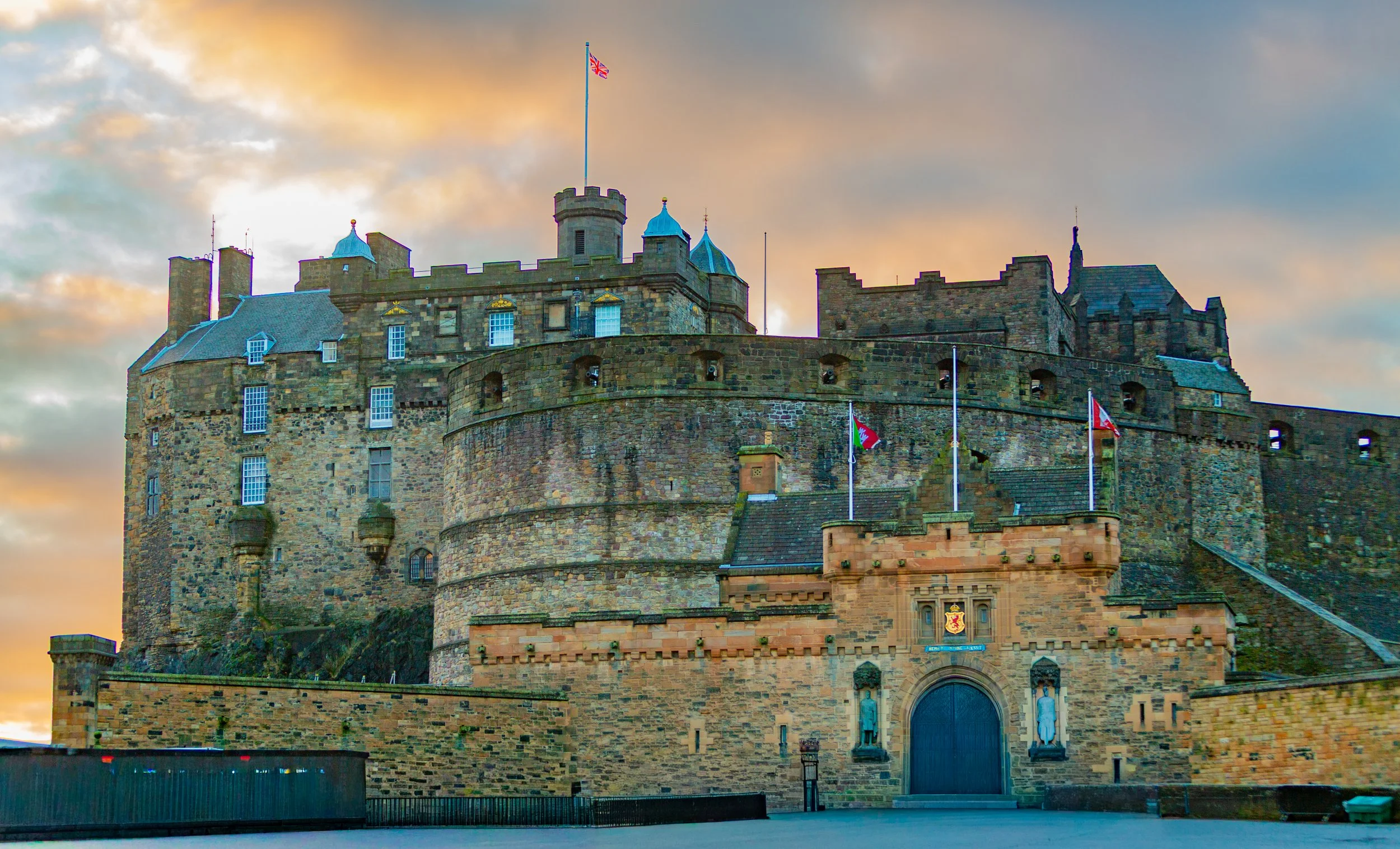 A historic stone castle with blue-topped turrets, flags, and an entrance, set against a sky with colorful clouds.