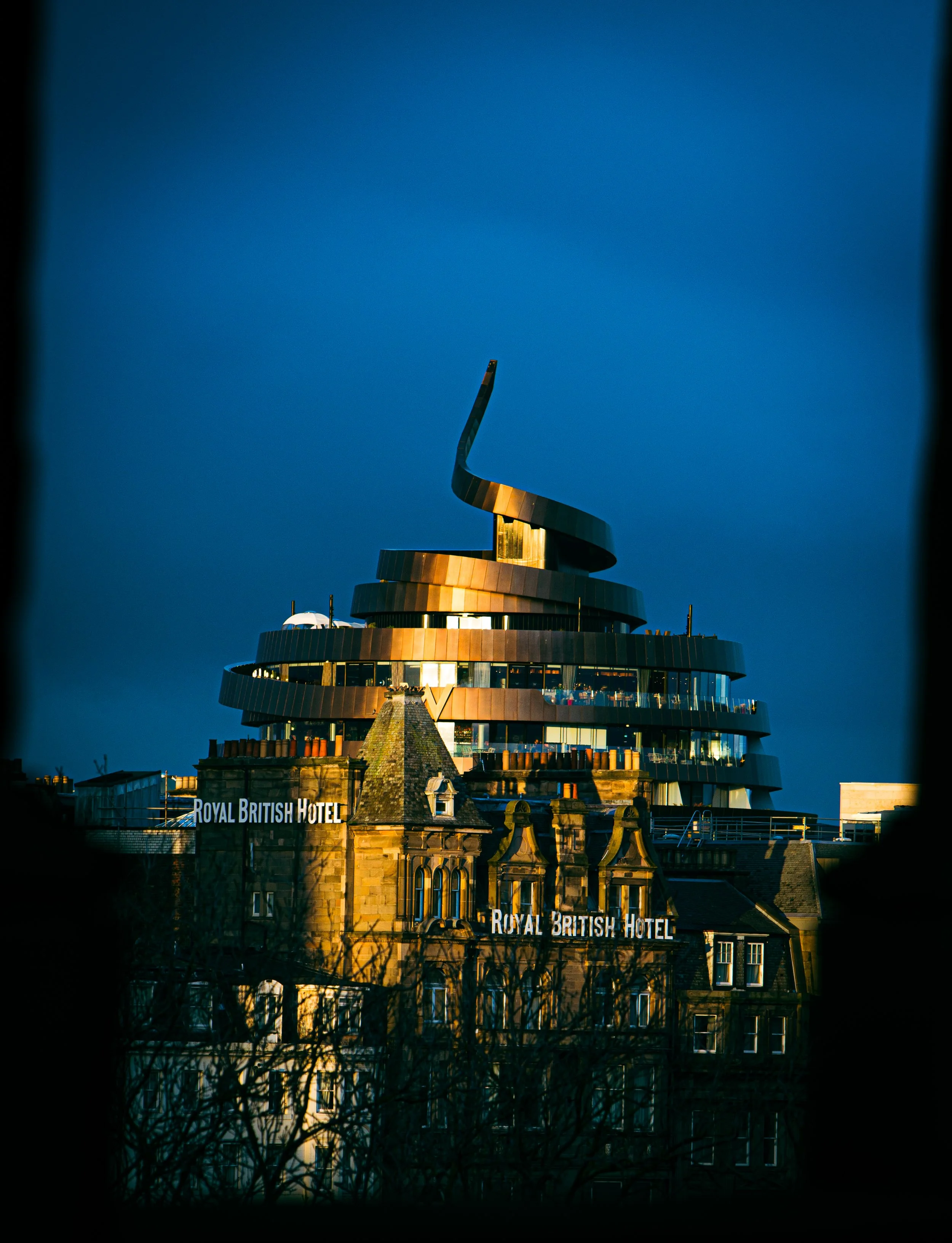 Nighttime view of the Royal British Hotel with a modern, spiral-shaped building illuminated against a dark blue sky. The building features prominent signs with the hotel's name and a historic stone structure at the base.