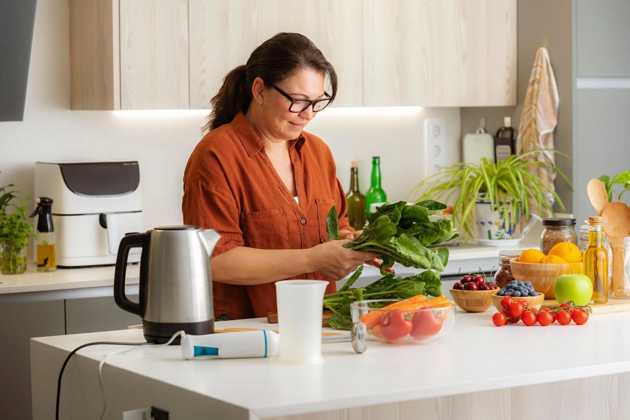 A woman with glasses and long dark hair, wearing an orange shirt, preparing vegetables in a bright kitchen with light wood cabinets and numerous fresh produce on the counter.