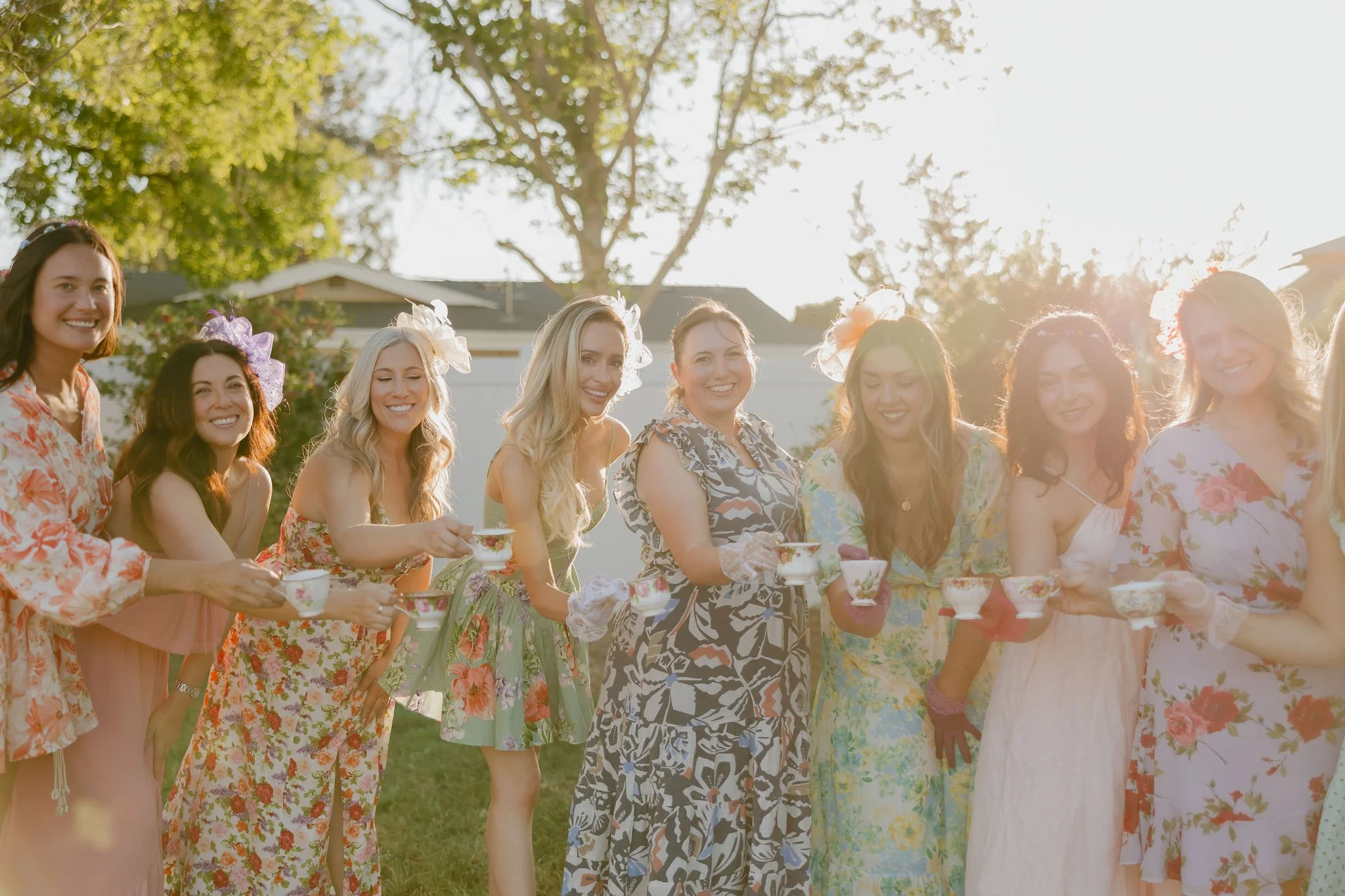 A group of women in floral dresses standing outdoors during a celebration, holding teacups, with trees and houses in the background, under bright sunlight.