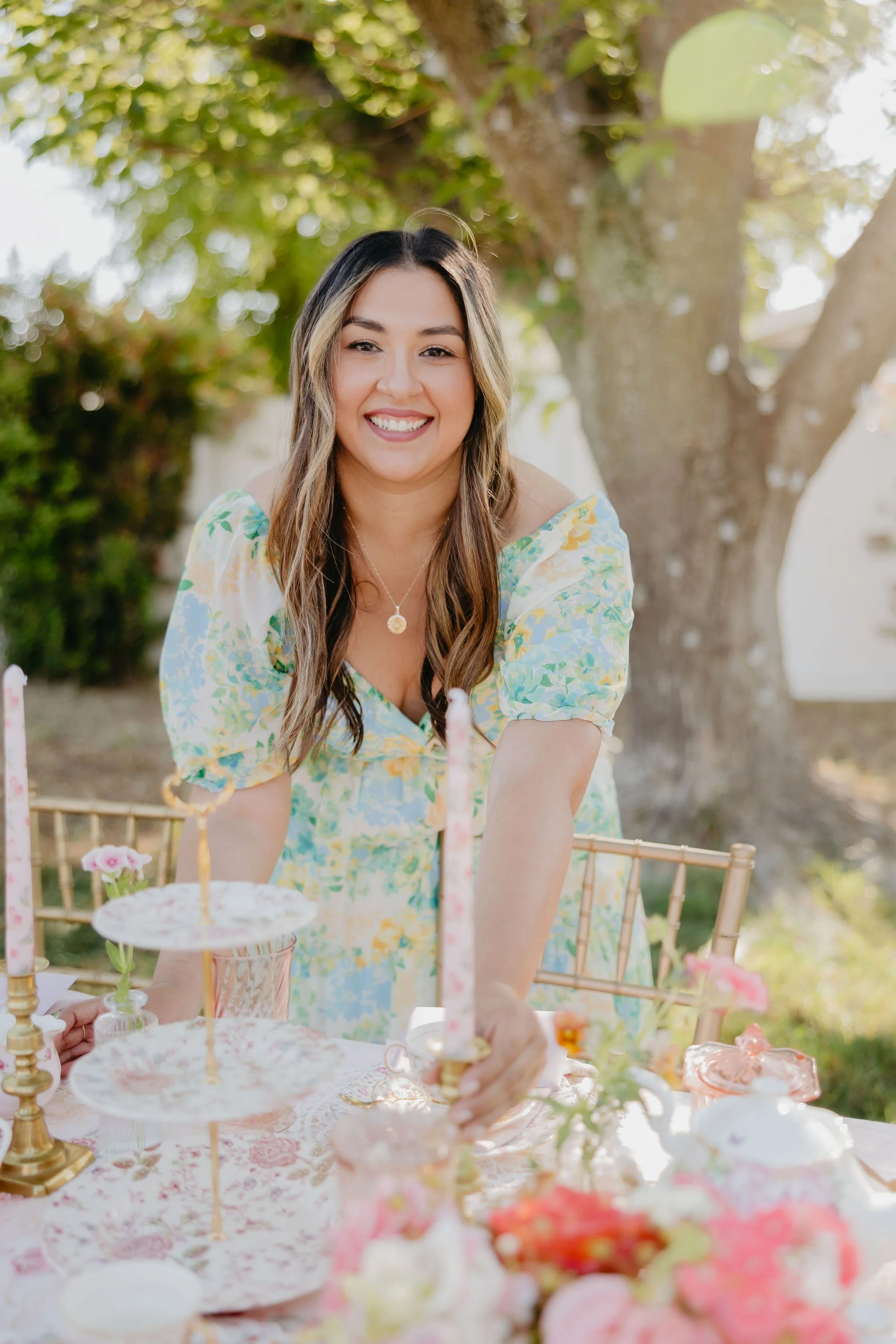 Woman smiling at a dessert table outdoors during daytime, with pink and white desserts and floral decorations.