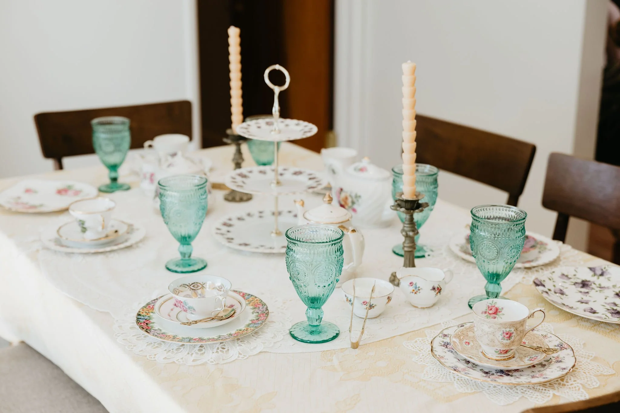 A dining table set with vintage china teacups, saucers, and tall green glass goblets, with a tiered dessert stand and candlesticks with taper candles.