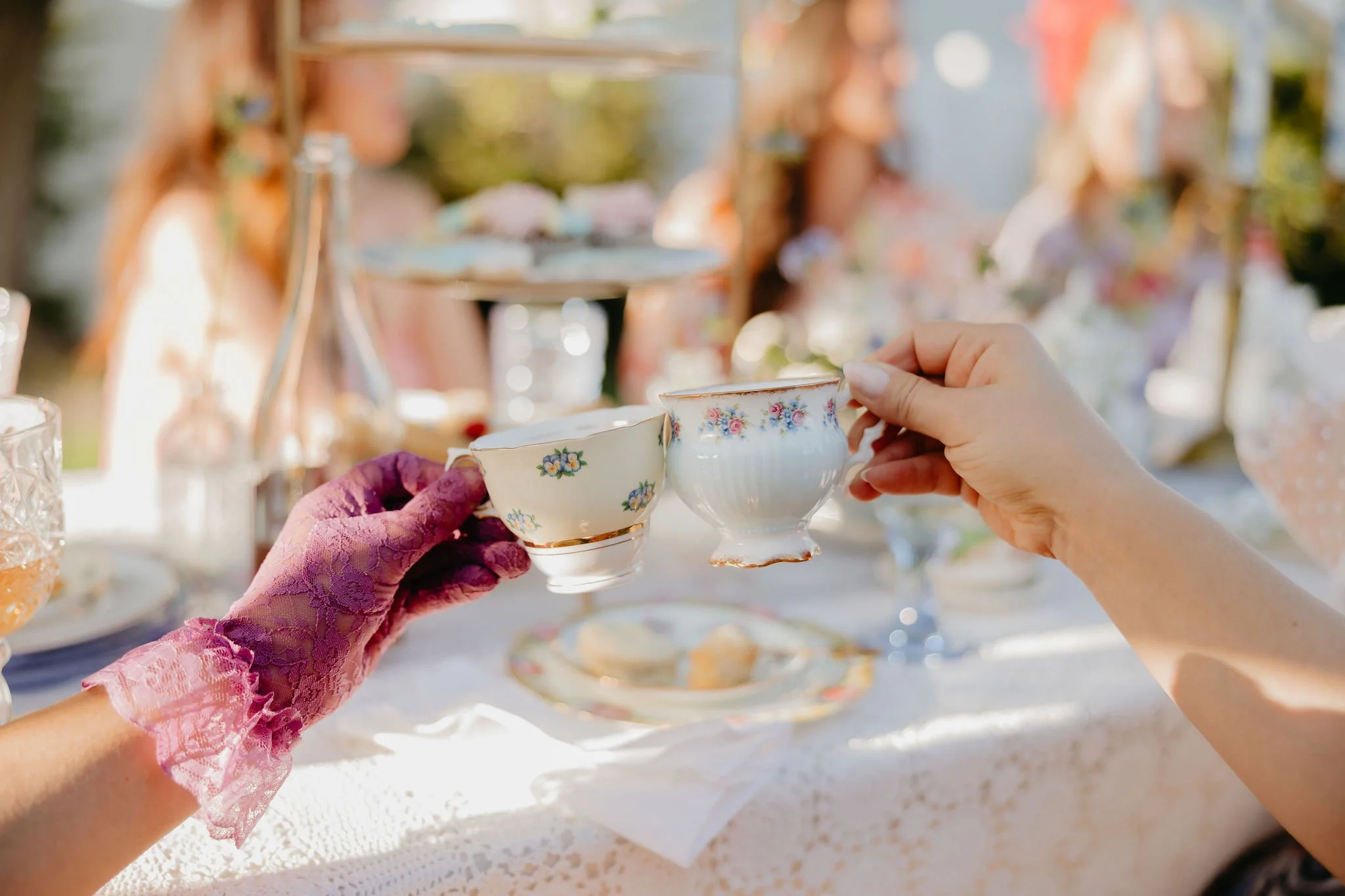 Hands raising vintage floral china teacups during a tea party outdoors, with a table set with plates and glasses, under bright natural light.