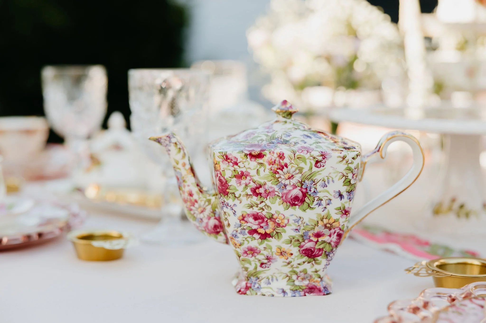 A floral-patterned teapot at a table set with china and gold-trimmed dishes, likely during a tea party or similar event.