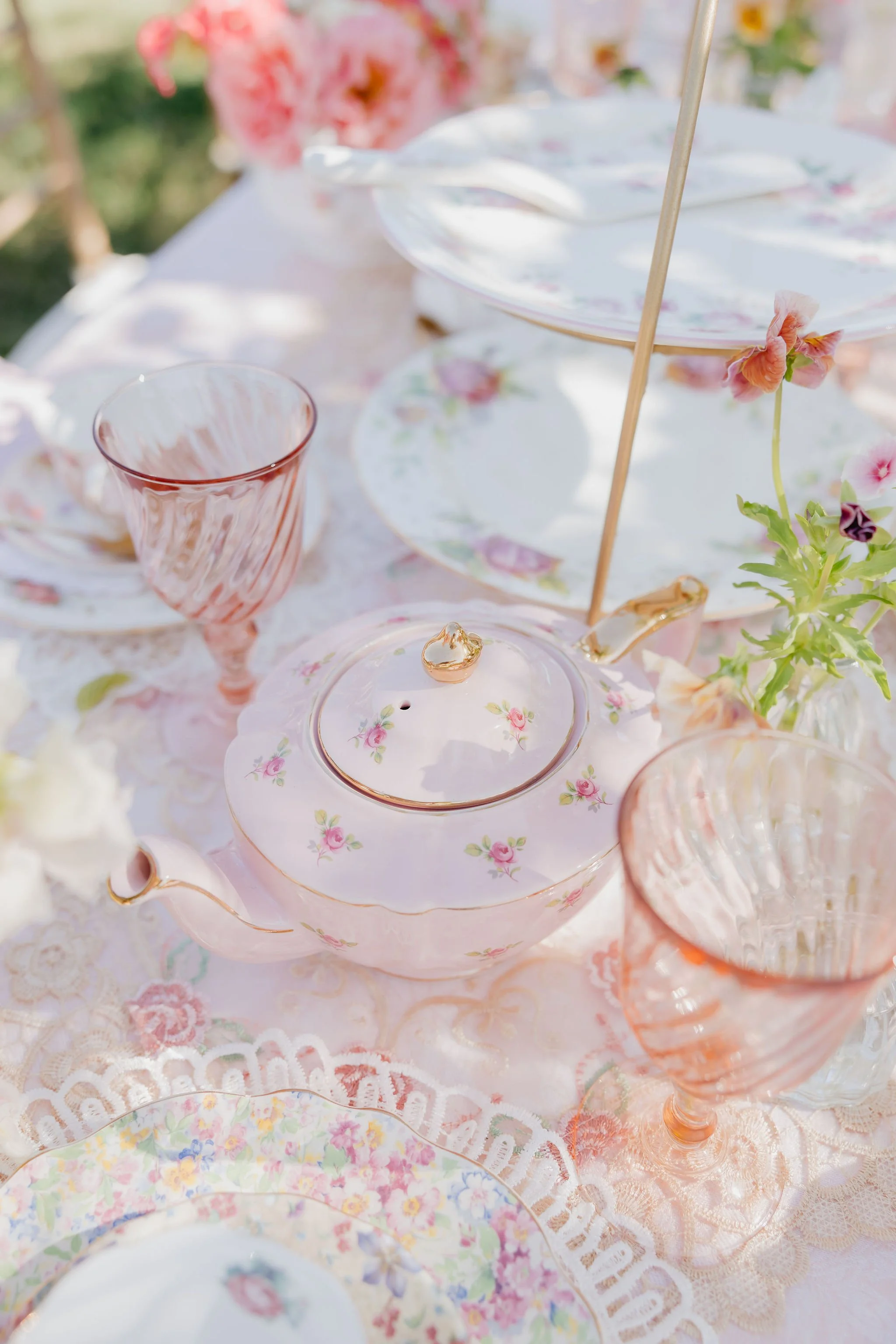 A pastel-colored tea set with floral patterns, pink glasses, and a lace tablecloth, accompanied by pink and white flowers, arranged outdoors for a tea party