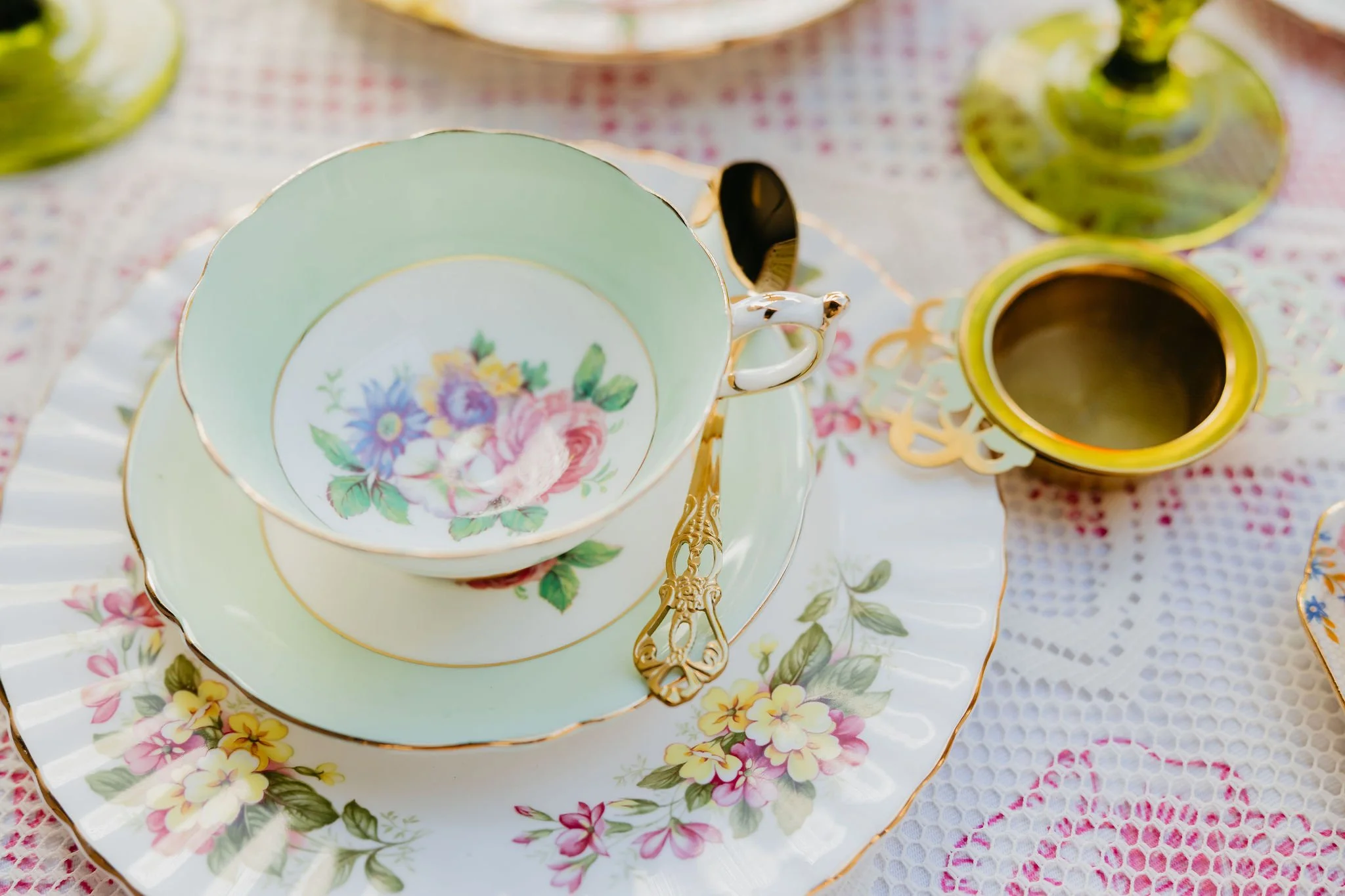 A set of vintage floral china tea cups and saucers with gold trim, decorated with colorful flower patterns, sitting on a floral tablecloth.