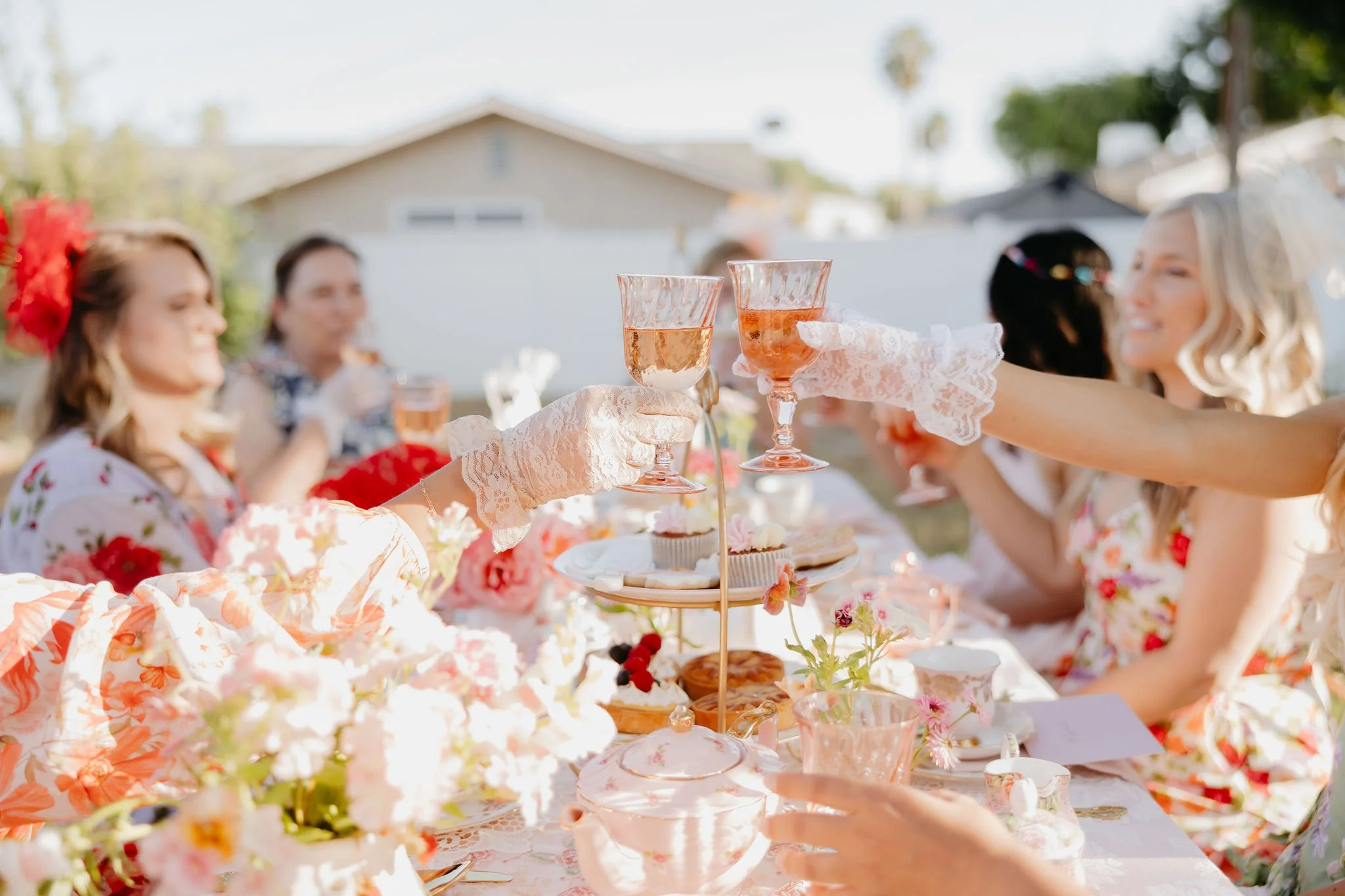 People raising glasses in a toast at a garden party with tableware, flowers, and desserts