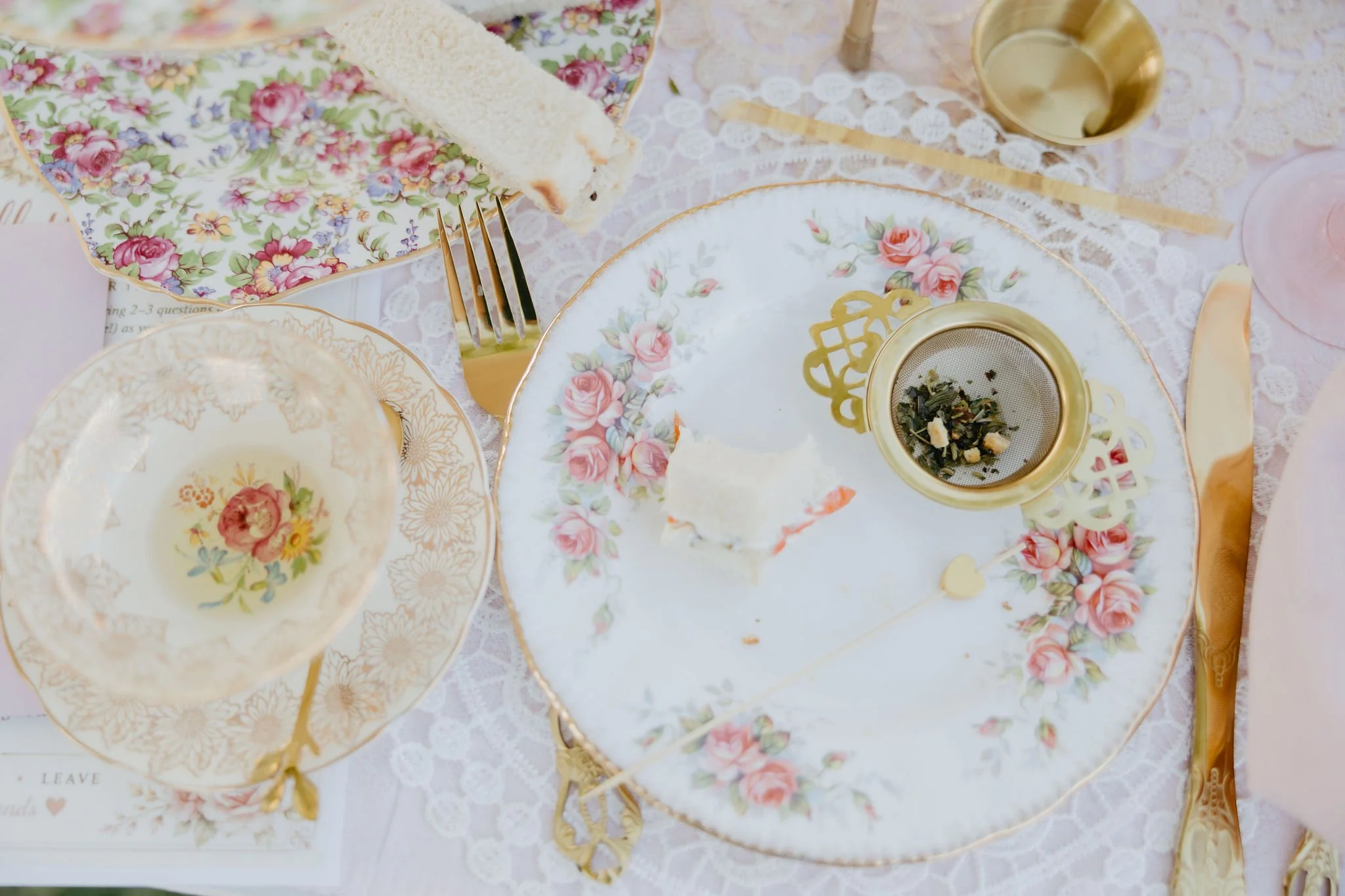 A wedding or formal event table setting with floral patterned china, gold accents, and a lace tablecloth. There is a piece of cake on a plate, a fork, and a gold heart-shaped cup or bowl.
