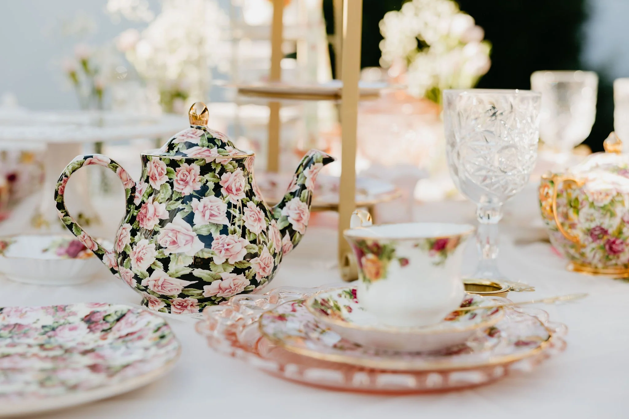 A floral patterned teapot and matching teacup set on a table with other glassware and tableware, with a blurred background of a bright, outdoor setting.