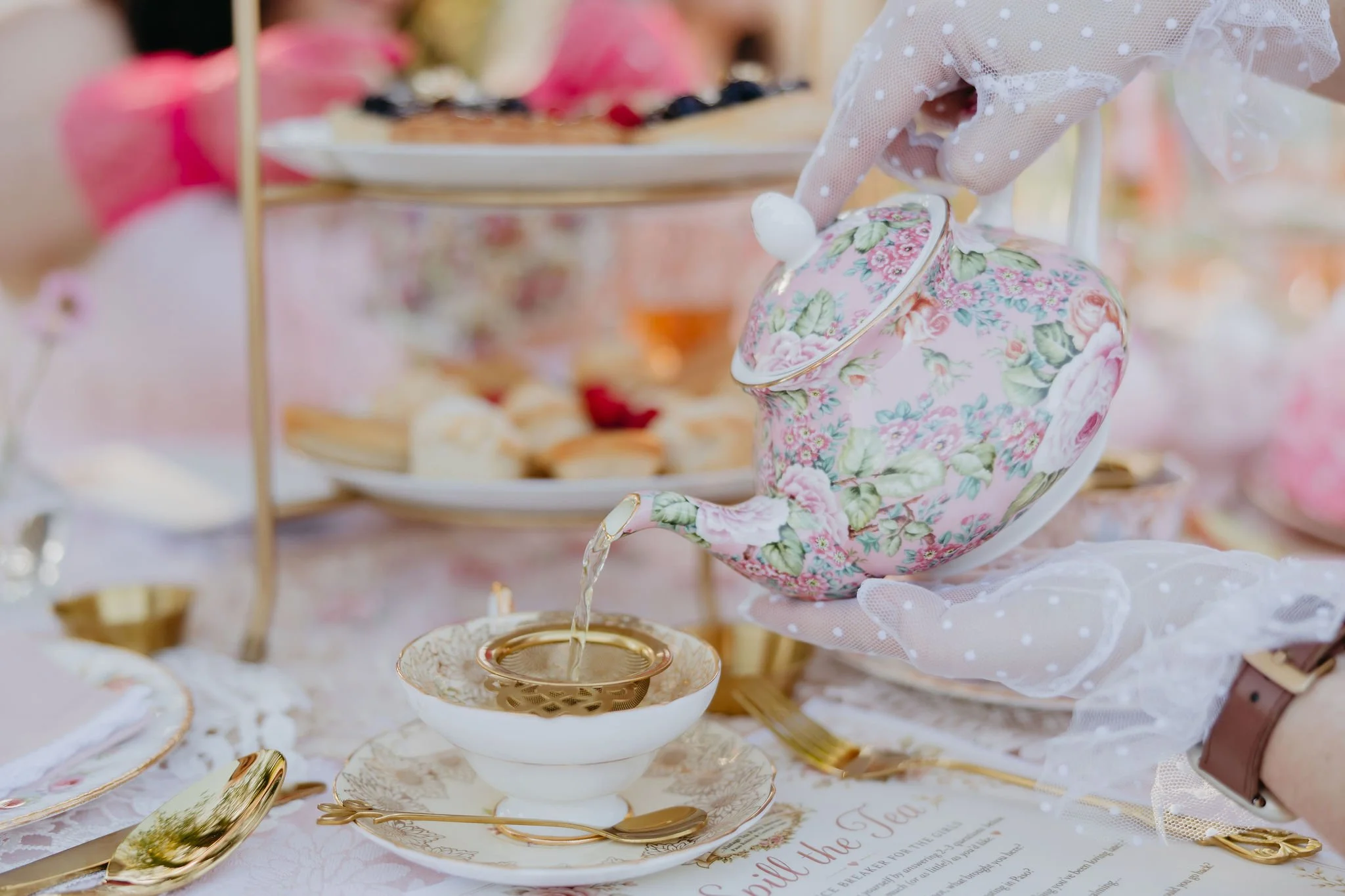A pastel-colored tea party setup with a floral teapot, teacup, and gold utensils on a table with a lace tablecloth. The background features a tiered tray with assorted desserts and pink decorations.