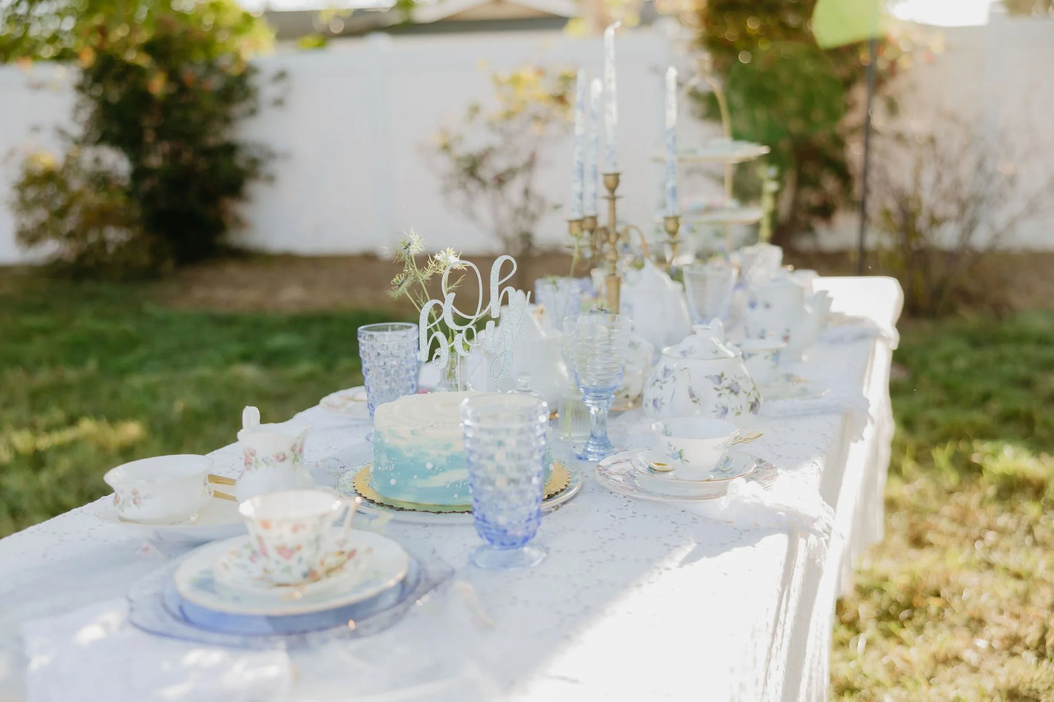 An outdoor table set for a celebration, decorated with a white tablecloth, several glassware, floral china teacups, and a blue and white cake through the middle. In the background, a white wall or fence and greenery are visible.