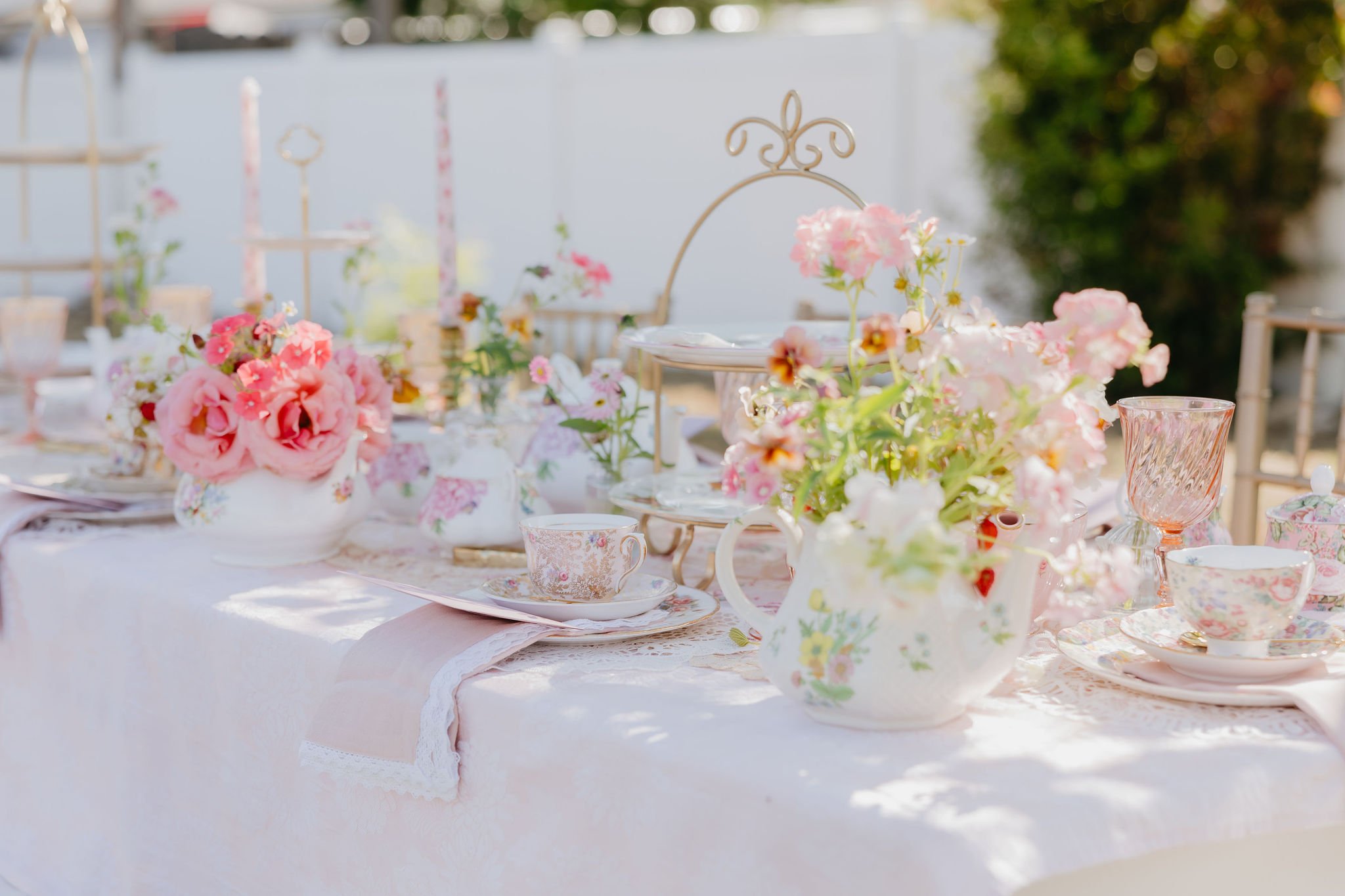 An outdoor table set for a celebration with pink flowers, china teacups, and a lace tablecloth, surrounded by greenery.