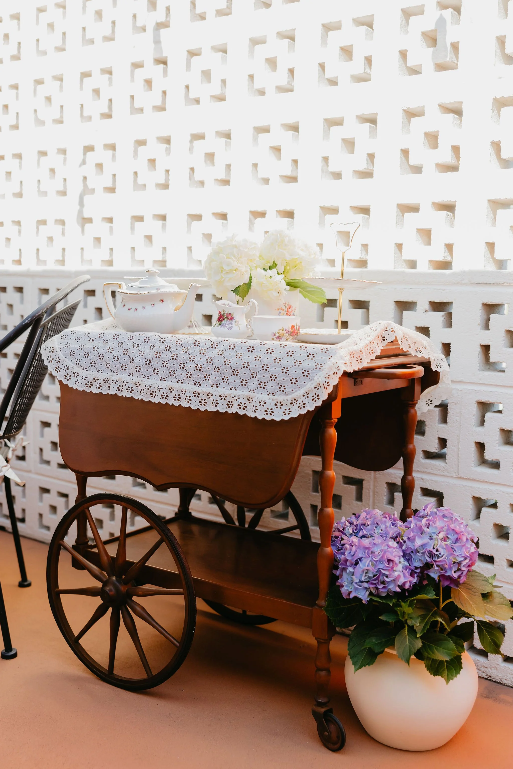 A vintage wooden cart with large wheels, a lace tablecloth, and a white teapot, cups, and a bouquet of white flowers on top. A potted purple hydrangea is on the ground beside it, set against a white lattice wall.