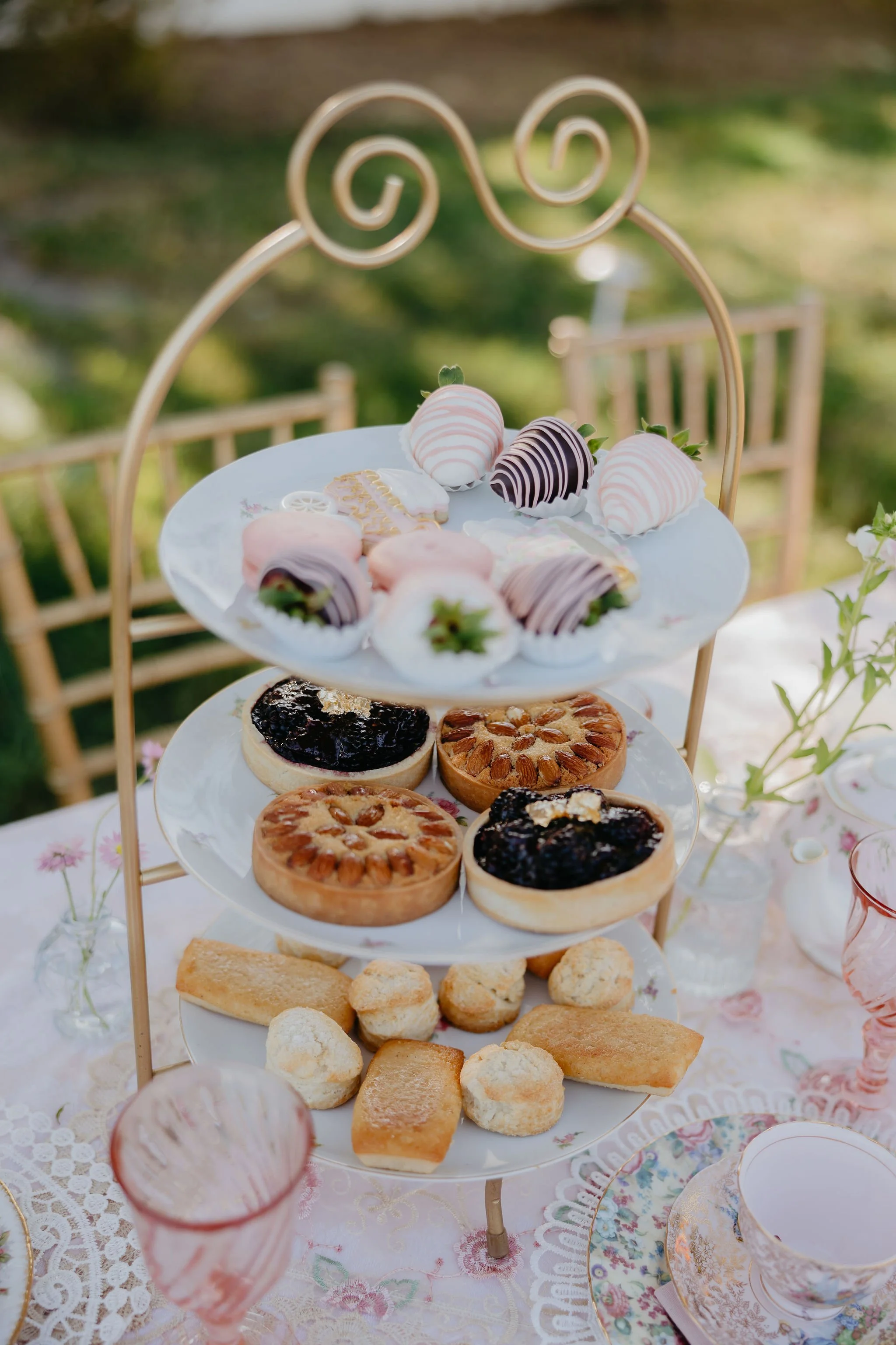Three-tiered serving tray with strawberries dipped in white and dark chocolate, assorted cookies, and finger sandwiches on a lace tablecloth, set outdoors with chairs and greenery in the background.