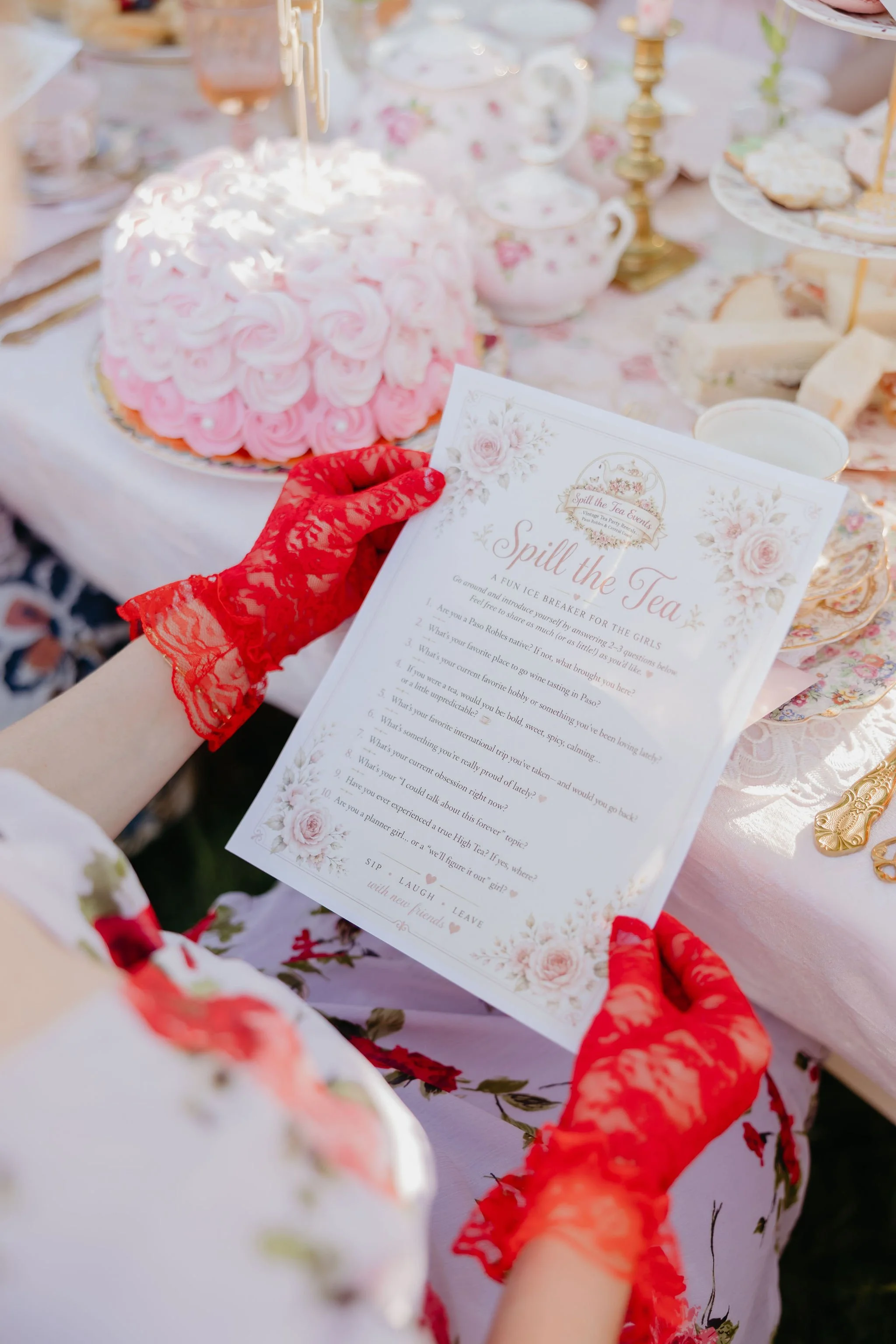 A wedding table setting with a pink rose-themed cake and vintage tea cups. A person wearing red lace gloves is holding a menu card titled 'Spill the Tea' with floral decorations.