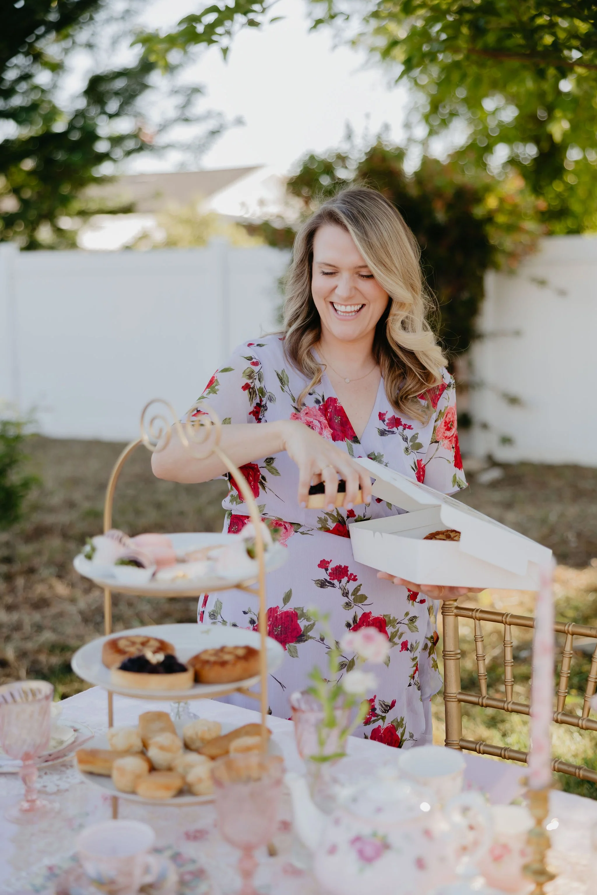 A woman with blonde hair in loose curls, wearing a white dress with red and green floral patterns, is smiling and holding a white box of cupcakes at an outdoor gathering. There is a table with desserts, including cookies and pastries, and a three-tiered stand with more baked goods. The setting includes trees, a white fence, and sunlight, indicating a garden or backyard party.