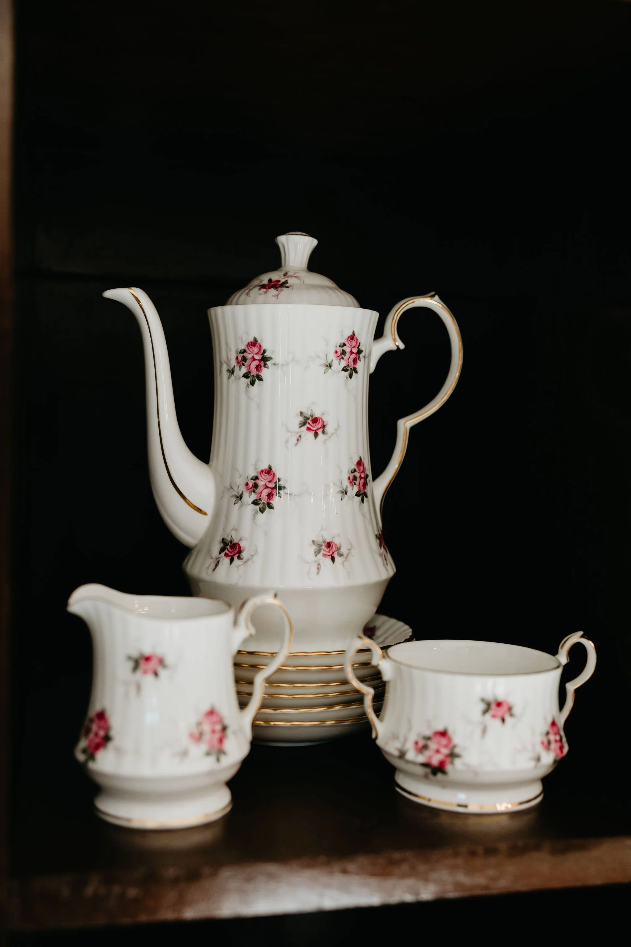 A set of white porcelain teapot and cups with pink roses and gold accents, displayed on a dark shelf.