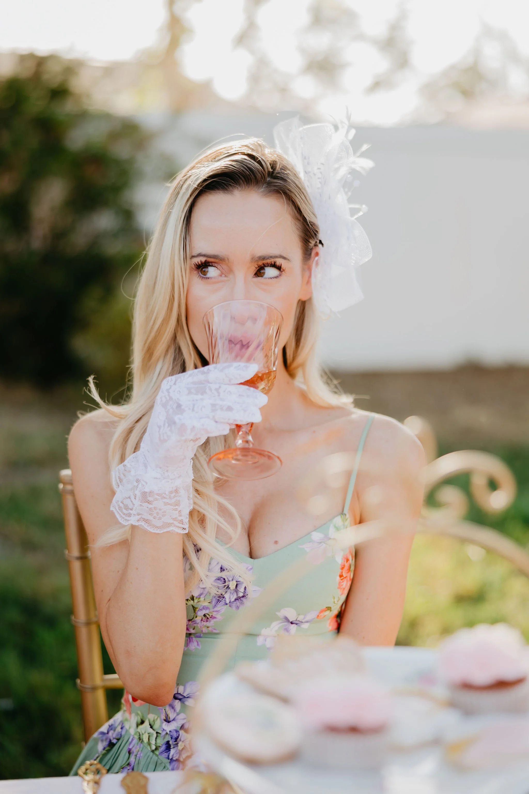 A young woman with blonde hair wearing a floral dress and white lace gloves, holding a wine glass and gazing to the side, at an outdoor gathering or tea party.