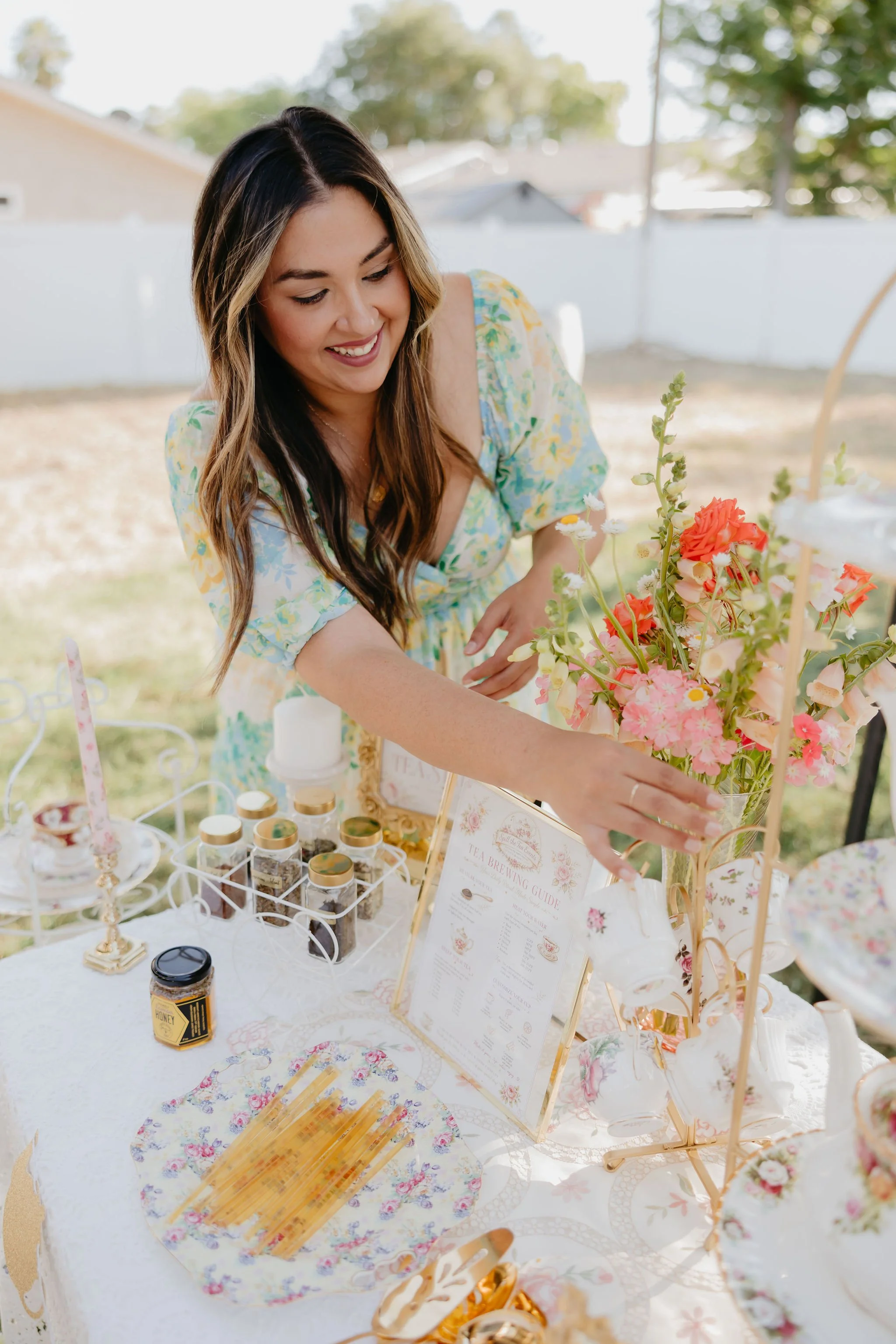 A woman arranging flowers at an outdoor table decorated for a celebration, with a floral & honey theme, including jars, a wine guide, and floral arrangements.