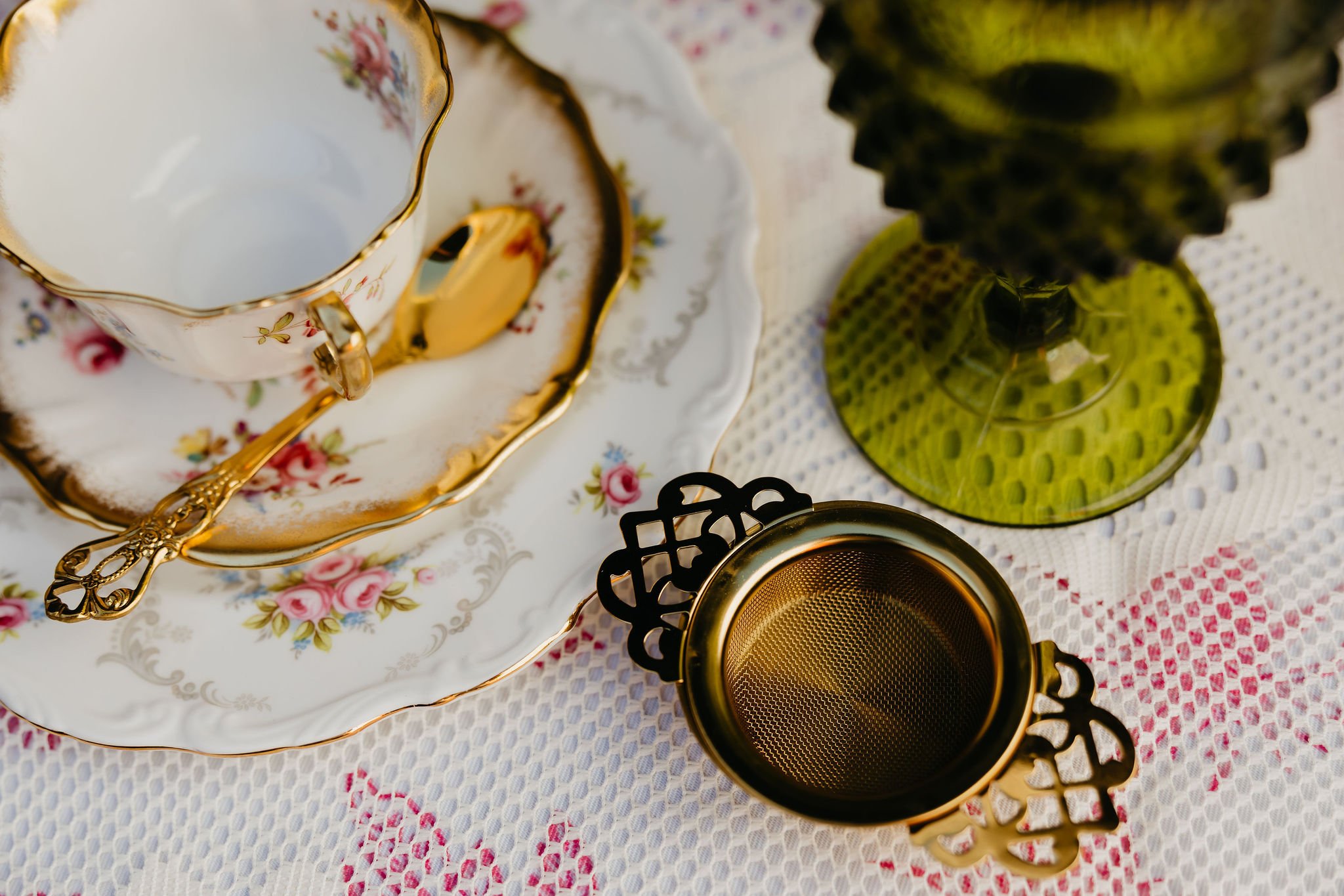 Vintage porcelain teacup and saucer with floral design and gold trim, a gold spoon, green glass candle holder, and a gold mesh tea strainer on a white lace tablecloth.
