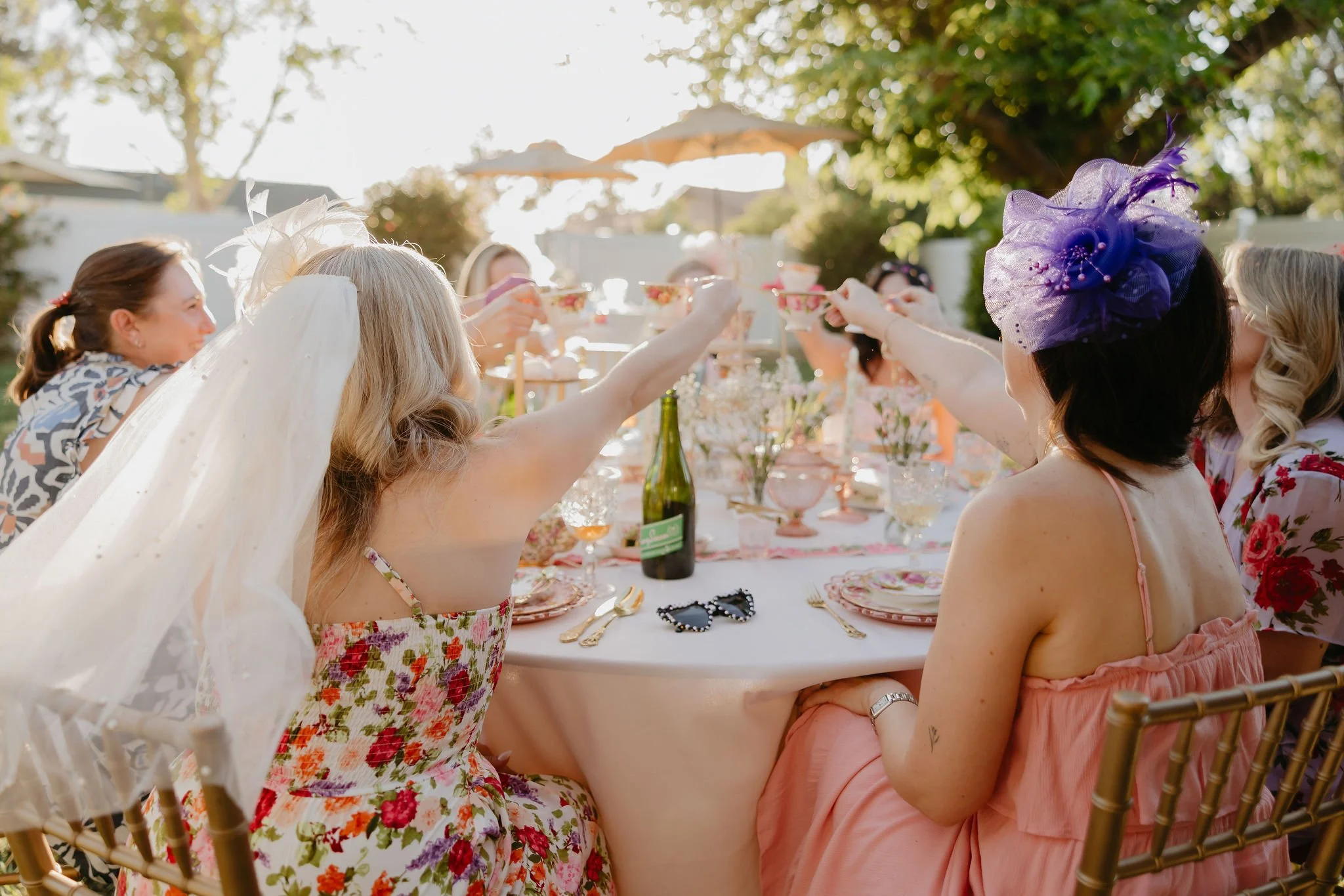 People gathered at an outdoor celebration raising their glasses in a toast, with a table set for a meal, in a garden with trees and umbrellas in the background.