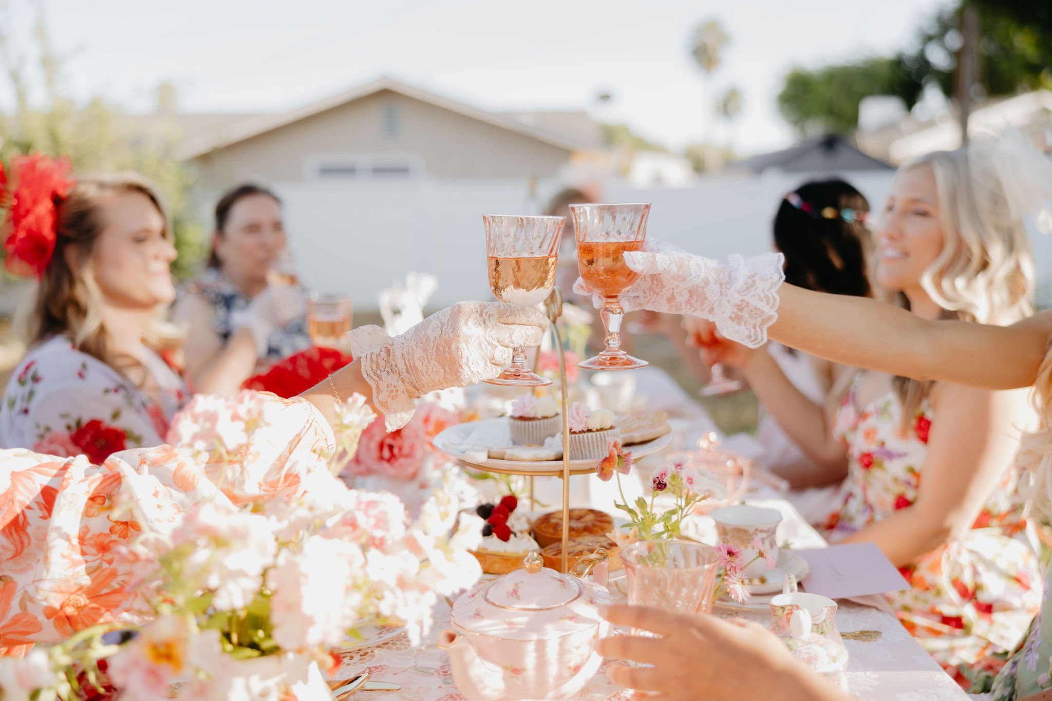 People at a party holding glasses of rosé wine, with a table of food and flowers outdoors.