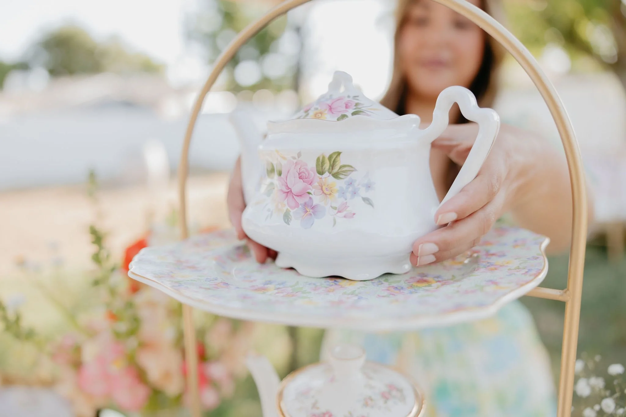 A woman arranging a white floral teapot on a tiered floral serving tray outdoors.