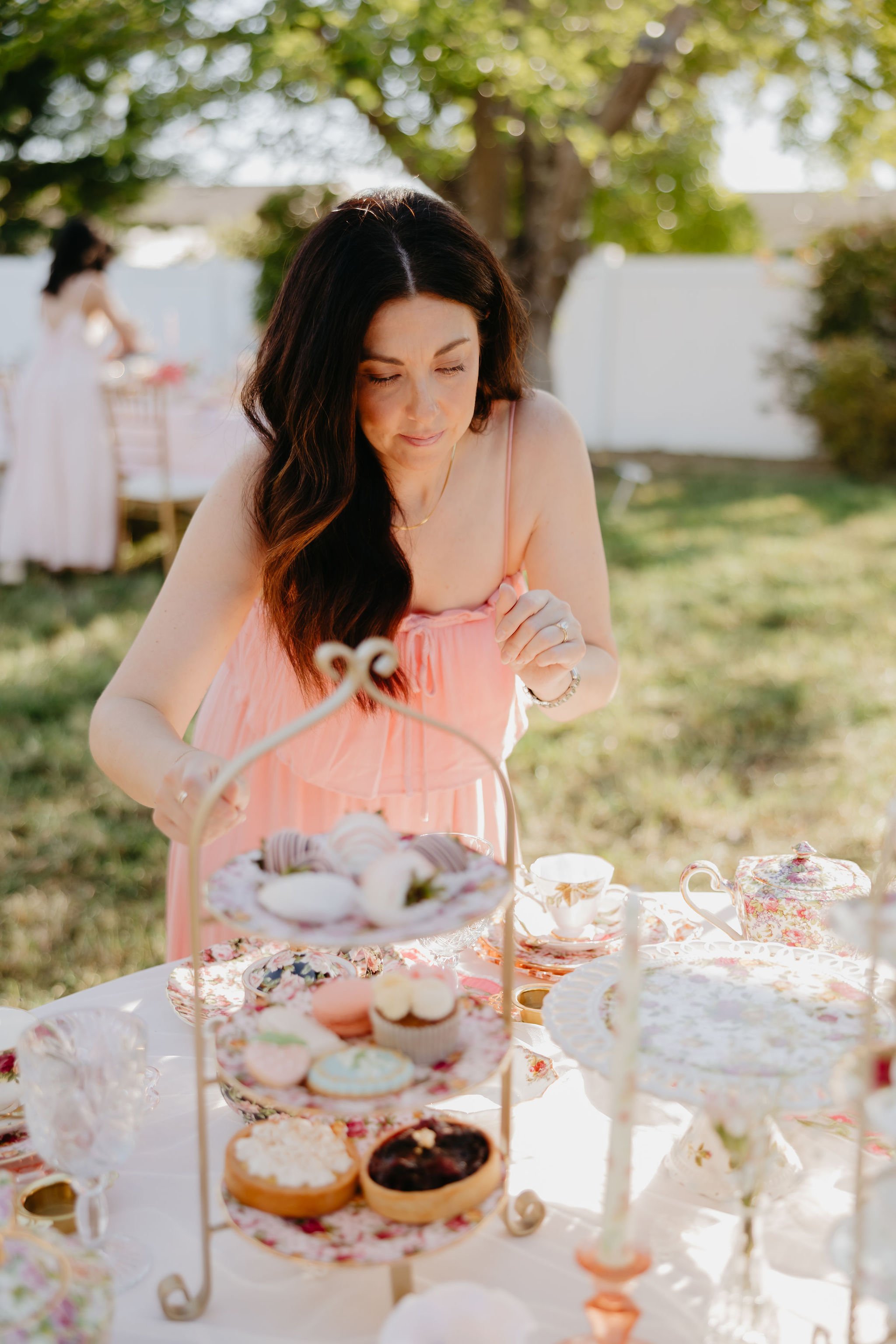 A woman with long dark hair in a pink dress is at an outdoor tea party table with a variety of desserts, set in a garden with trees and greenery in the background.