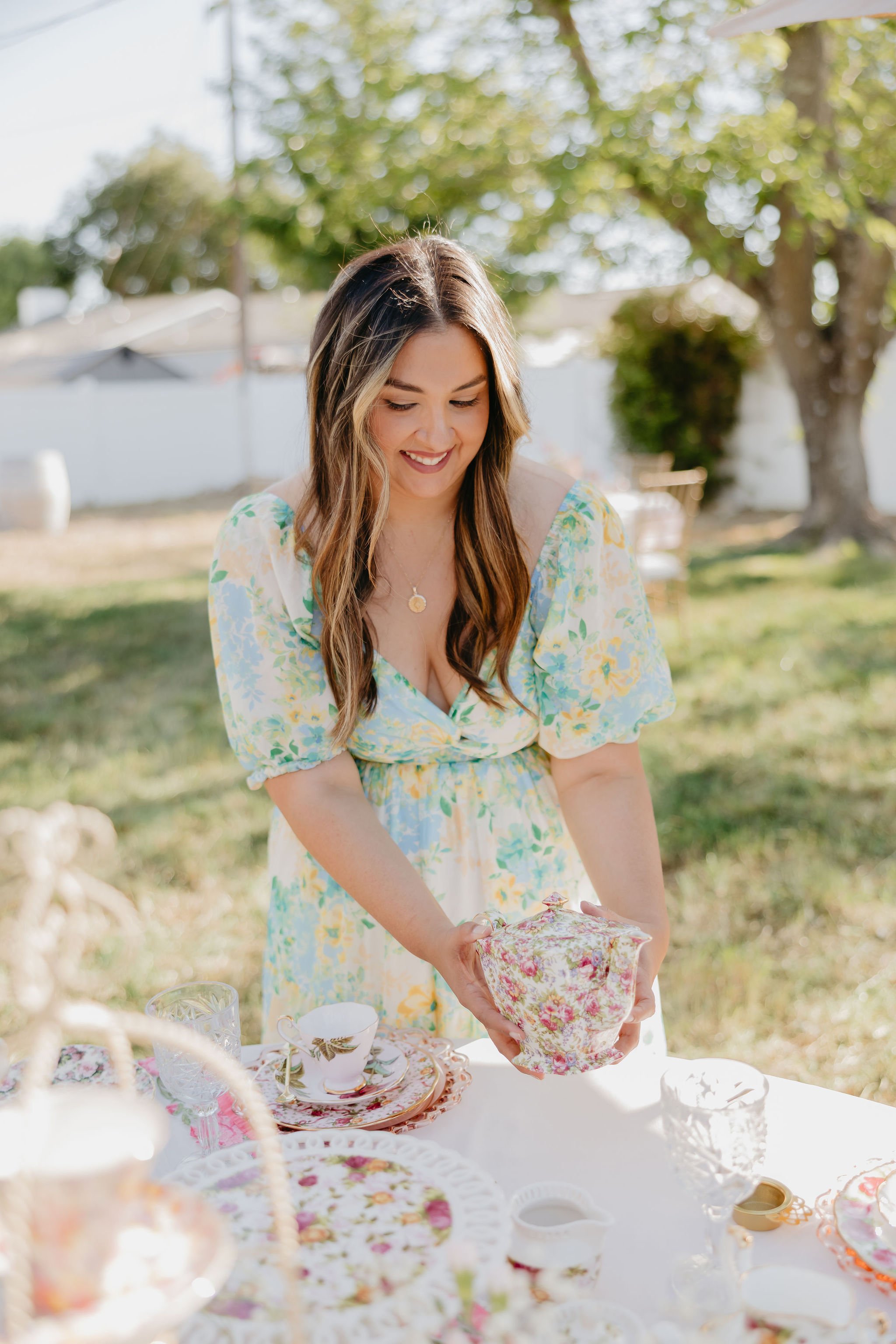 A woman in a pastel floral dress is setting a table with plate, cups, and teacups at an outdoor brunch or tea party on a sunny day, with trees and a white fence in the background.