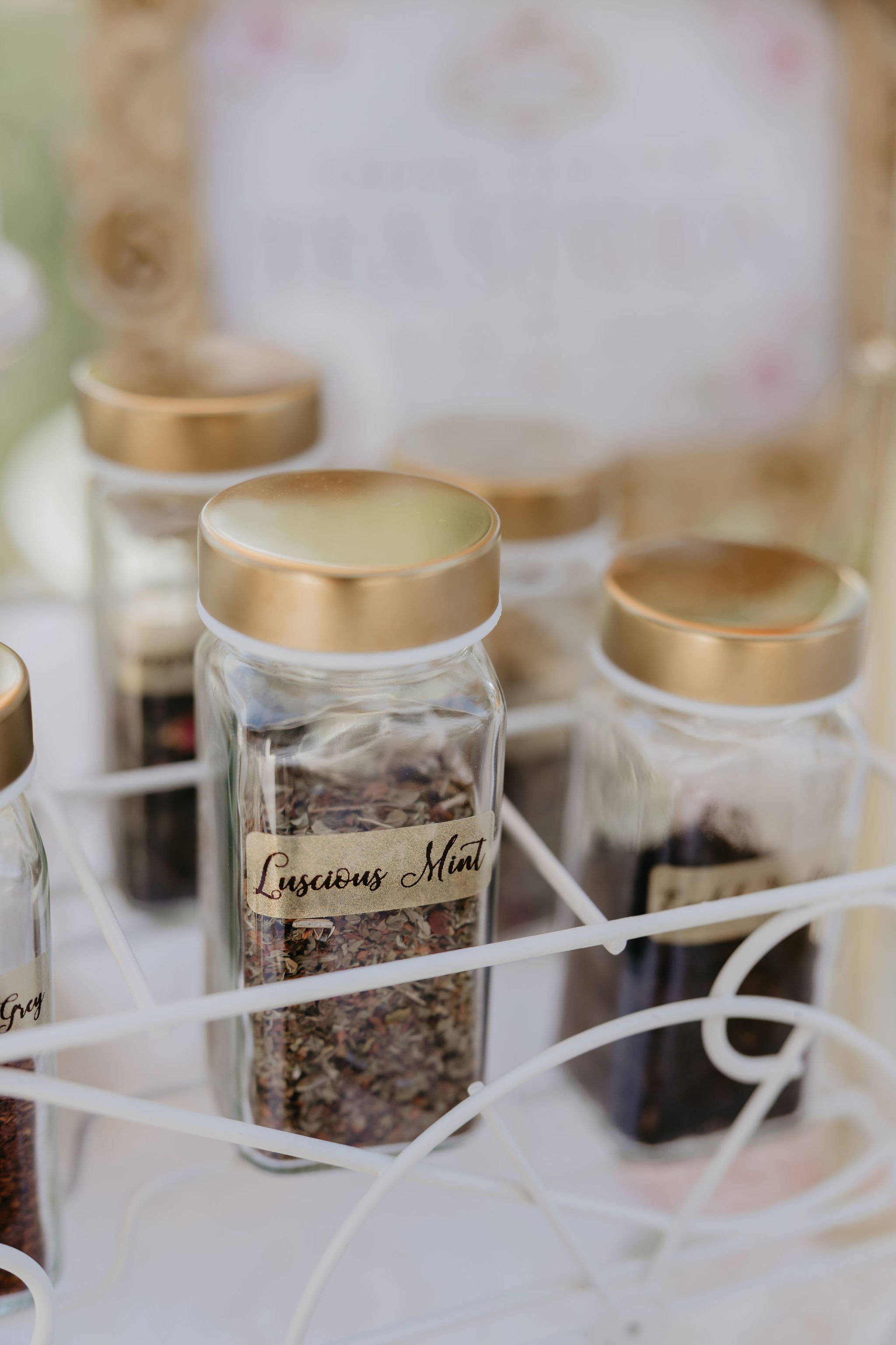 Close-up of small glass jars with gold lids, containing hand selected premium teas. One labeled "Luscious Mint," placed in a white wire rack.