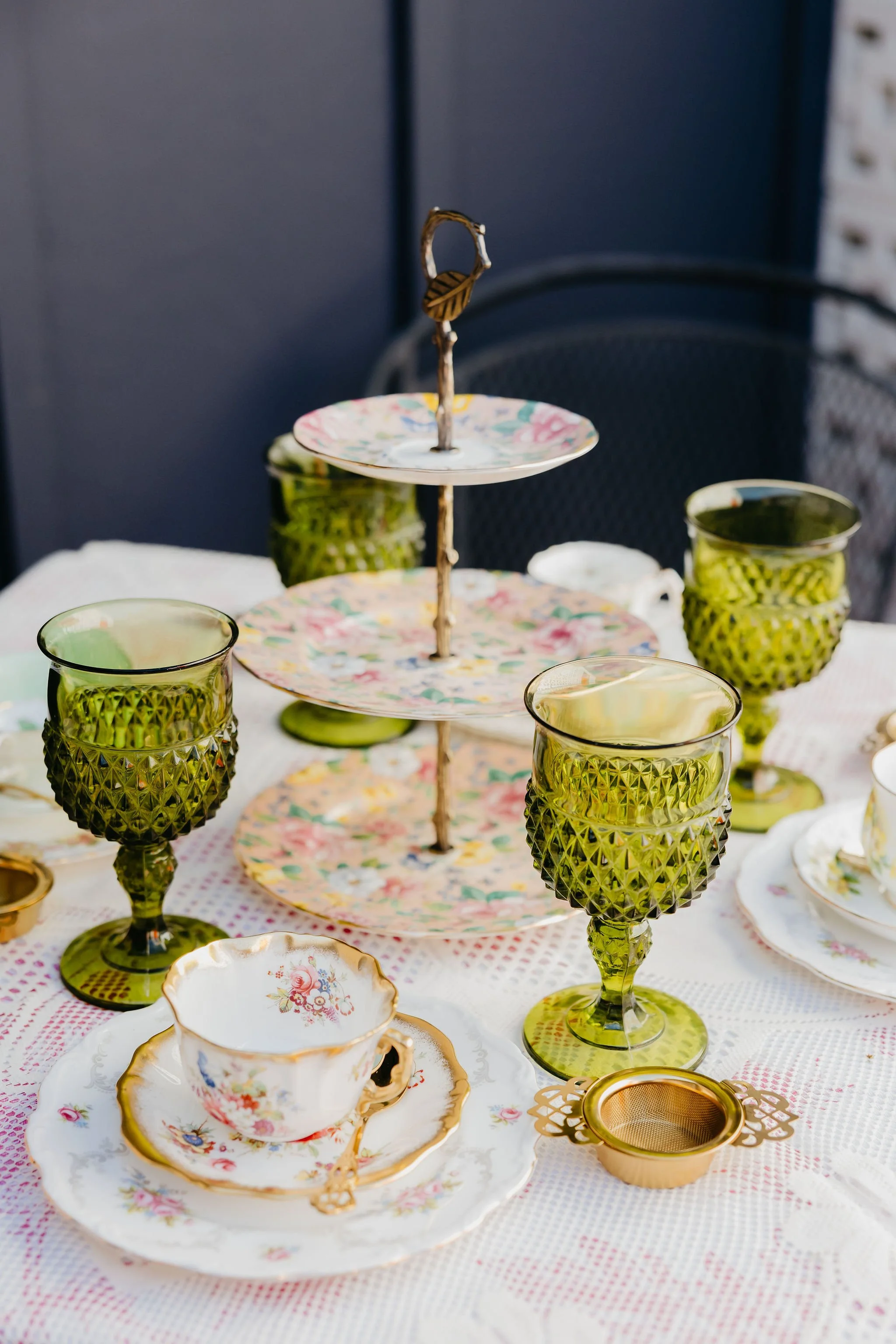 A table set with vintage teacups, plates, and green glassware, featuring a dessert stand with floral plates.