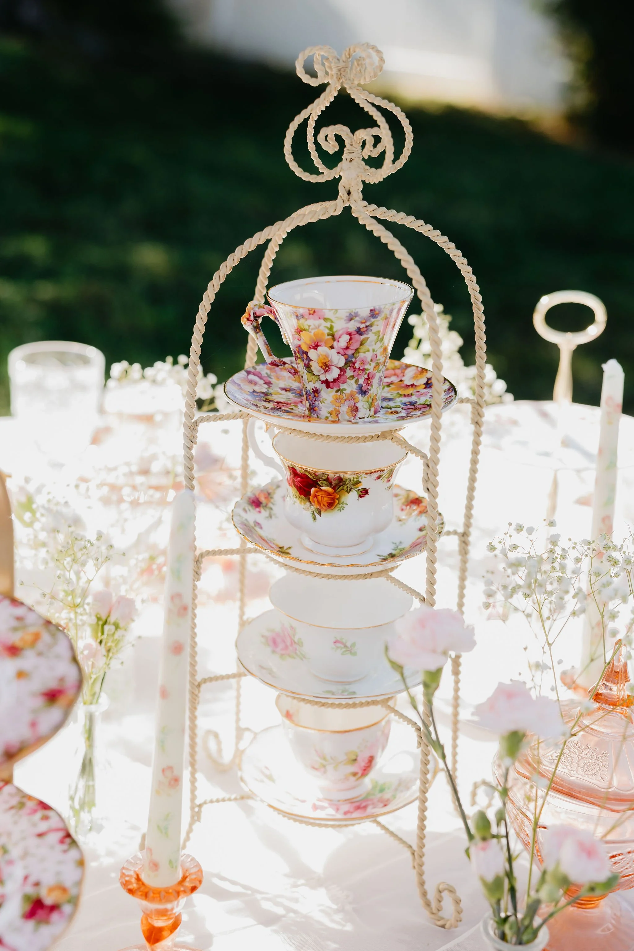 A tiered display of floral teacups and saucers on a white table, decorated with flowers, set outdoors with greenery in the background.